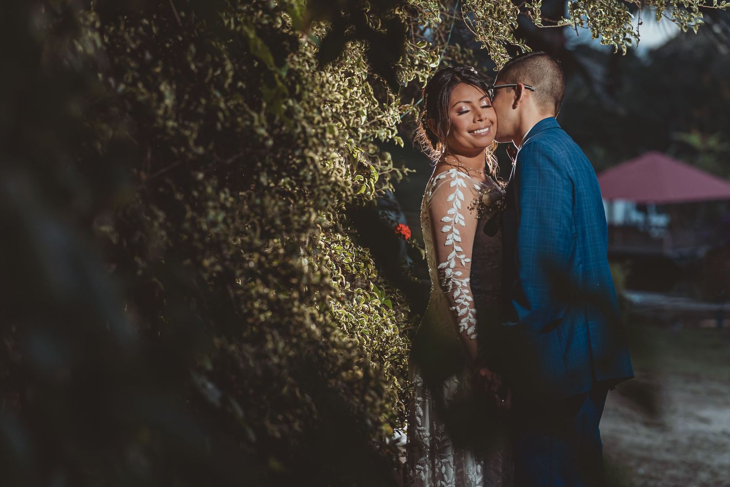 Novio (traje azul) besa cuello de novia sonriente (vestido manga encaje); foto boda exterior naturaleza.