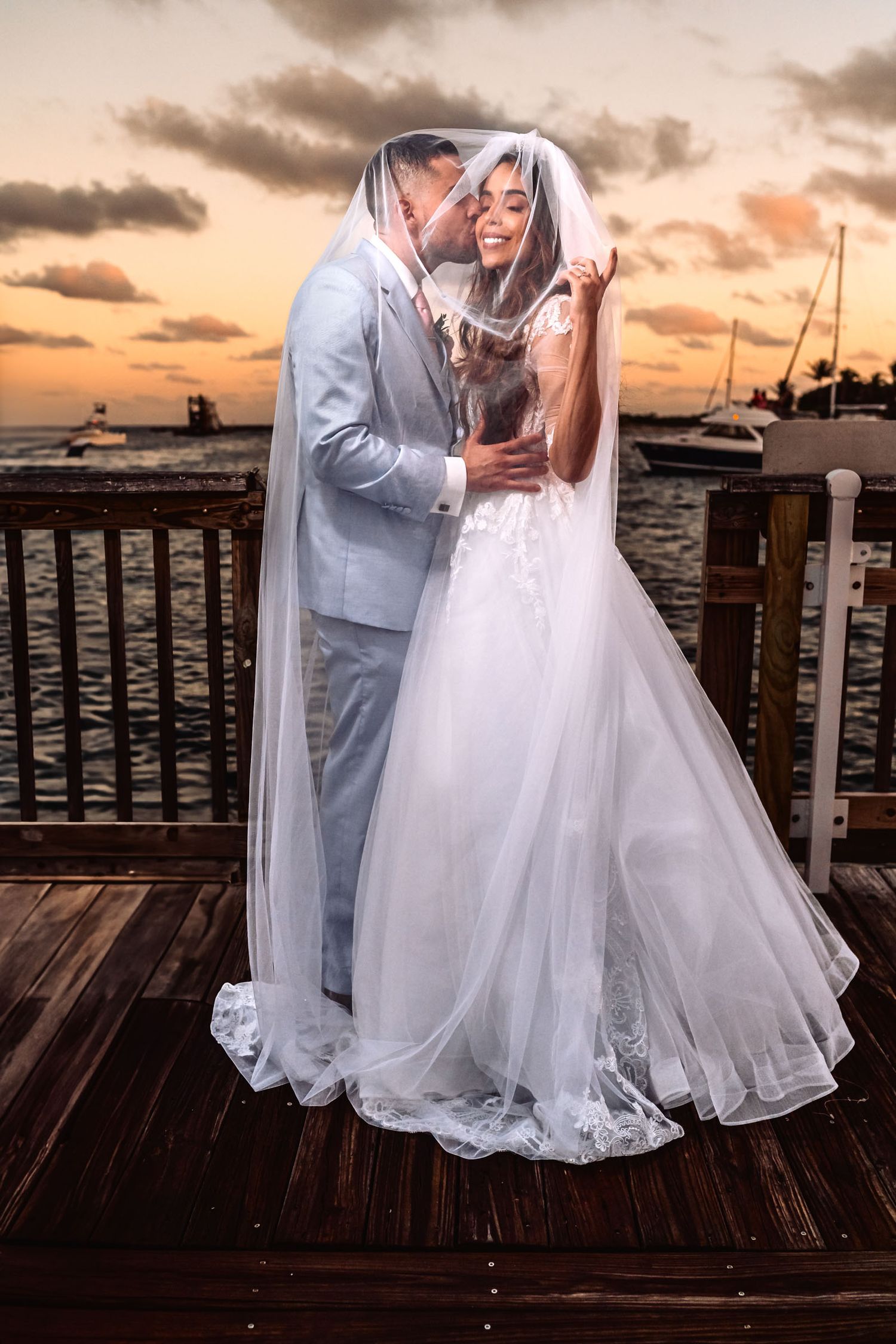 Pareja abrazada en boda al atardecer con velo ondeando. Cielo dramático con nubes y luz dorada. Siluetas románticas en paisaje natural