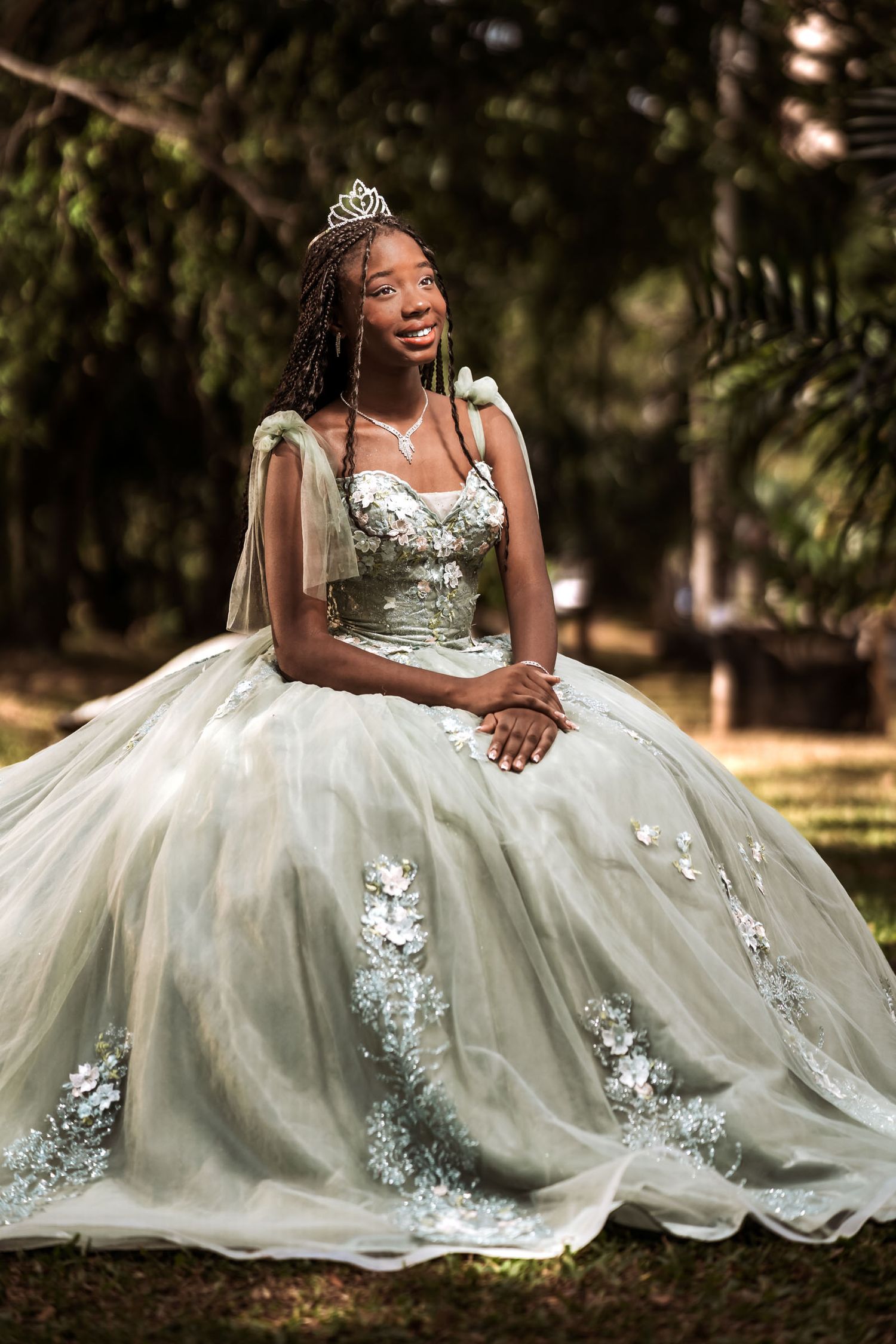 Quinceañera afrodescendiente con vestido princesa verde/plata y tiara, sentada sonriente en parque/bosque.