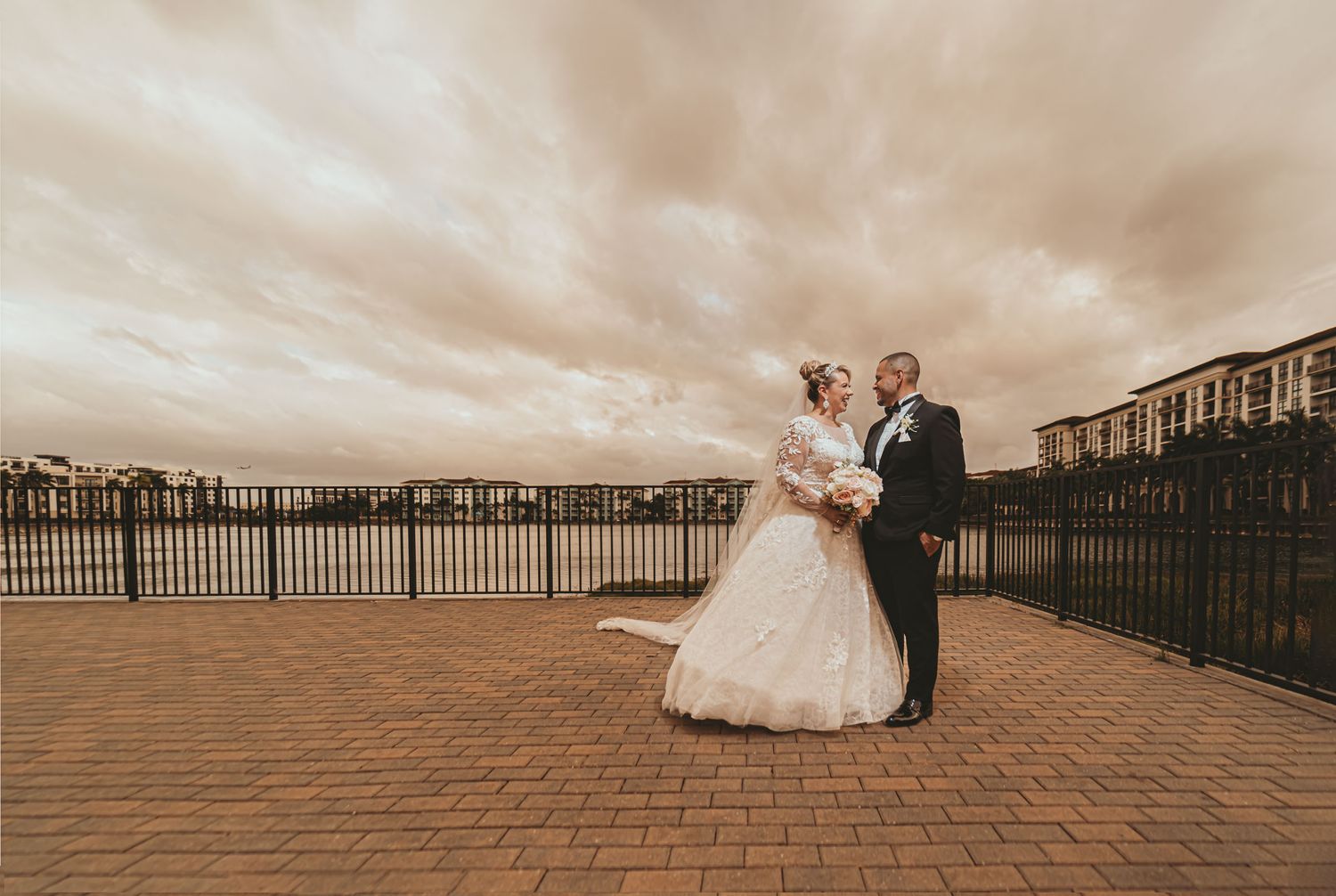 Novios se miran en terraza adoquinada junto a lago; cielo nublado dramático de Miami. Fotografía de boda exterior.