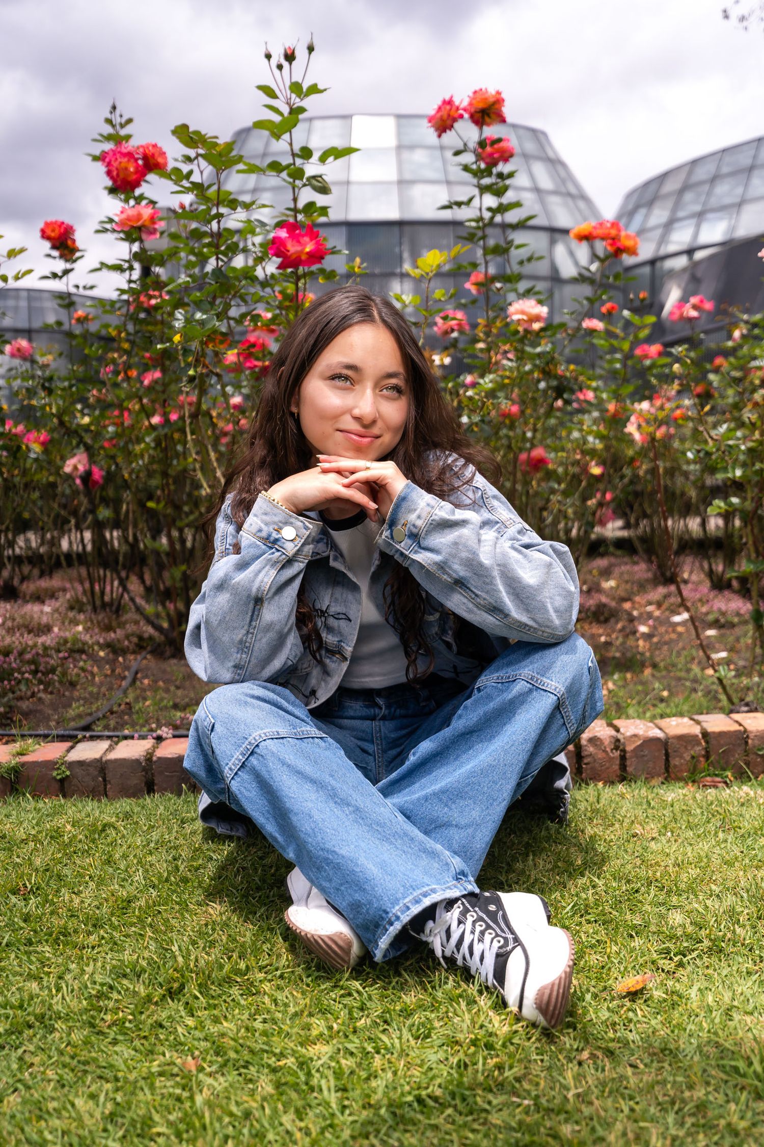 Joven en sesión prequince, chaqueta denim y jeans, sentada en césped ante rosales y edificio moderno.