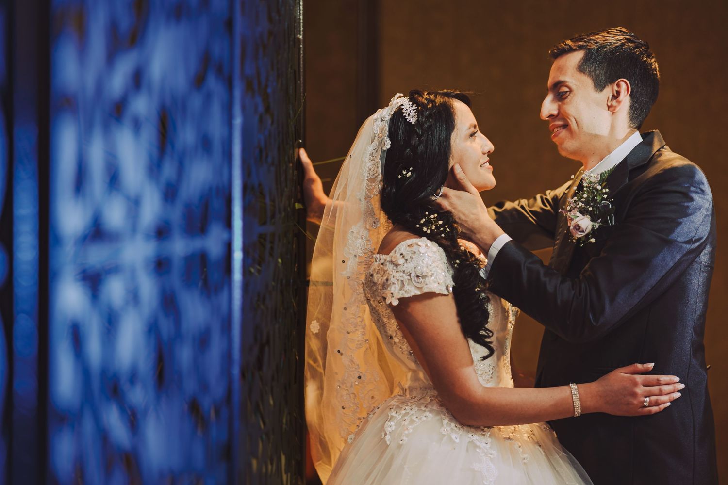 Novios se abrazan y miran sonrientes junto a pared azul; él acaricia su rostro. Boda romántica.