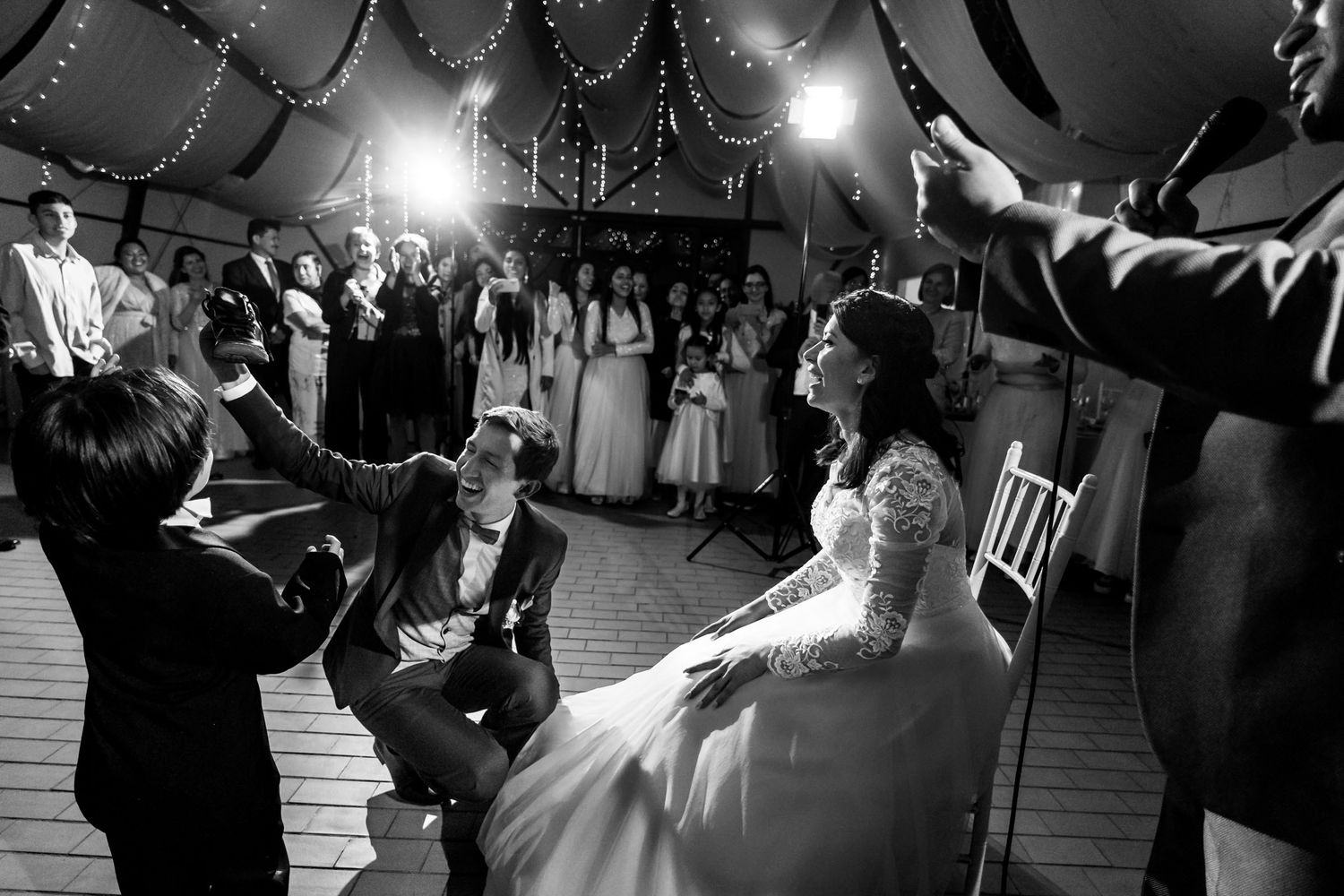 Recién casados en blanco y negro: Novio se arrodilla frente a la novia sentada, ambos ríen. Invitados observan en recepción.