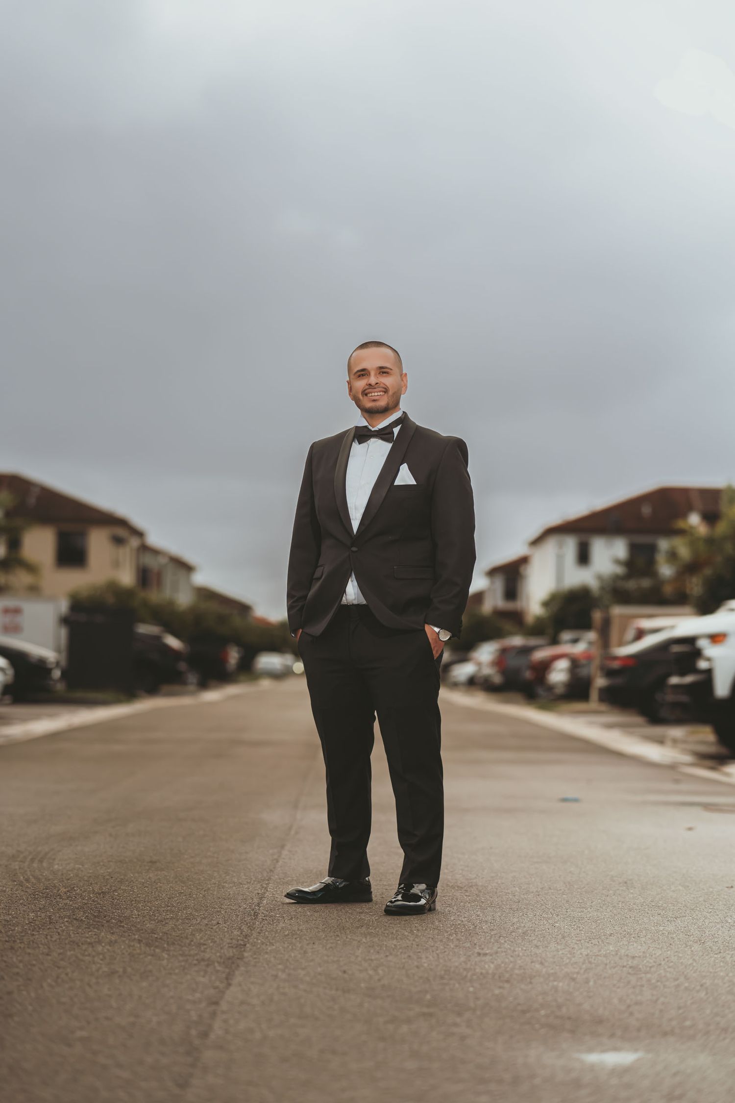 Novio elegante con esmoquin posa en medio de calle residencial bajo cielo nublado; retrato boda exterior en Miami.