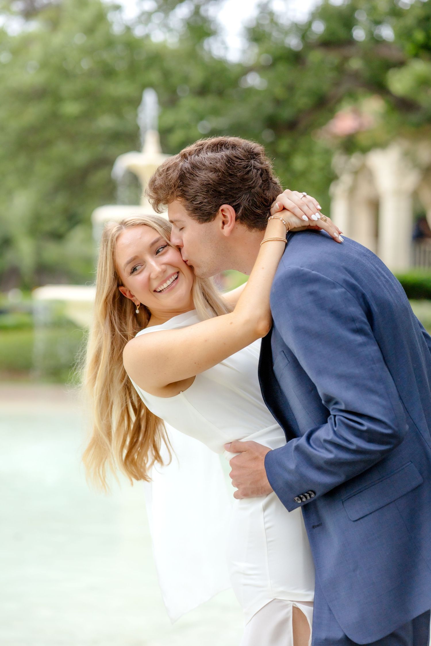 Bride-to-be in a white dress smiles as her fiancé kisses her cheek during their romantic engagement session at Flippen Park in Dallas, Texas