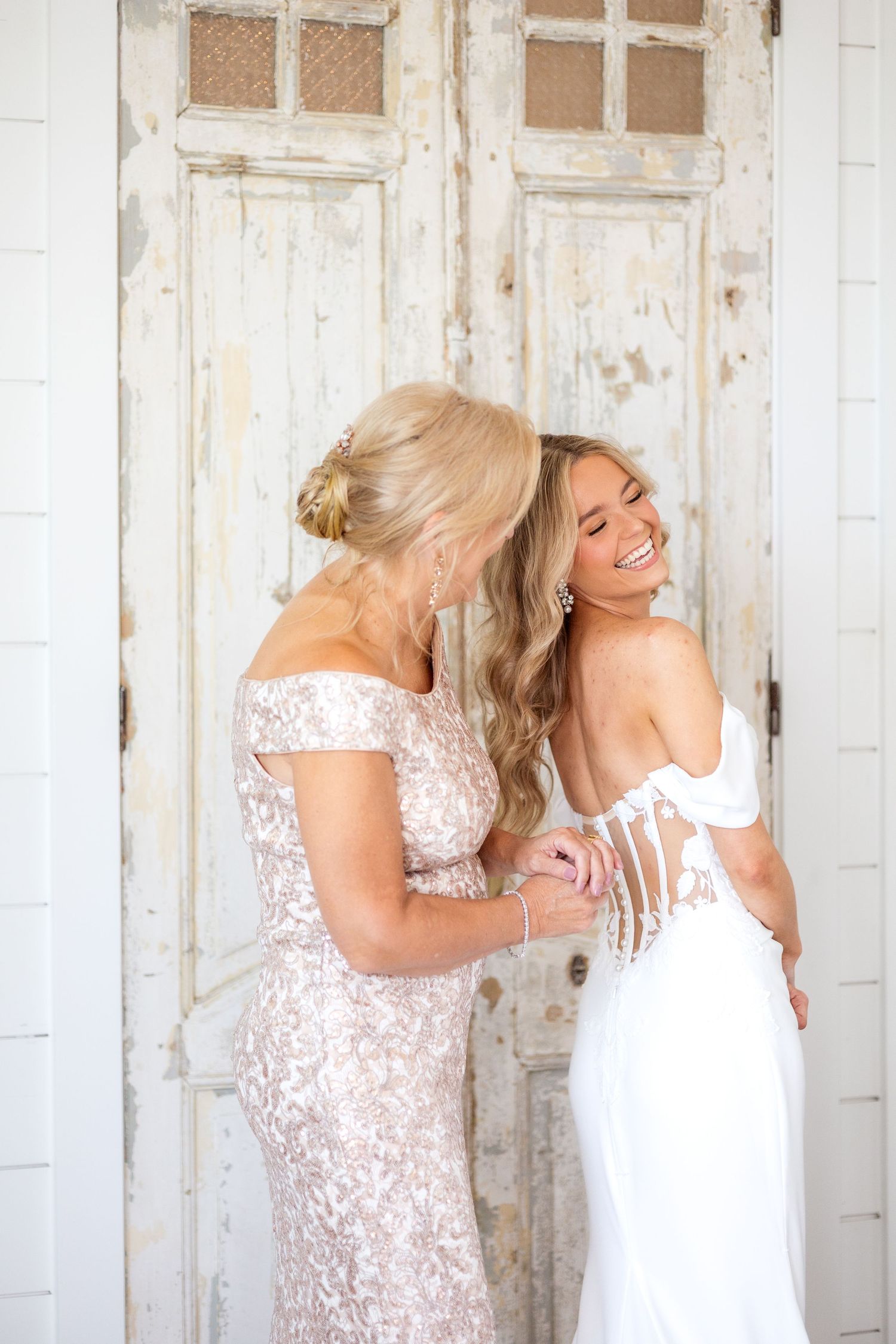 Mother buttoning the bride's wedding dress as the bride smiles toward her mother-in-law