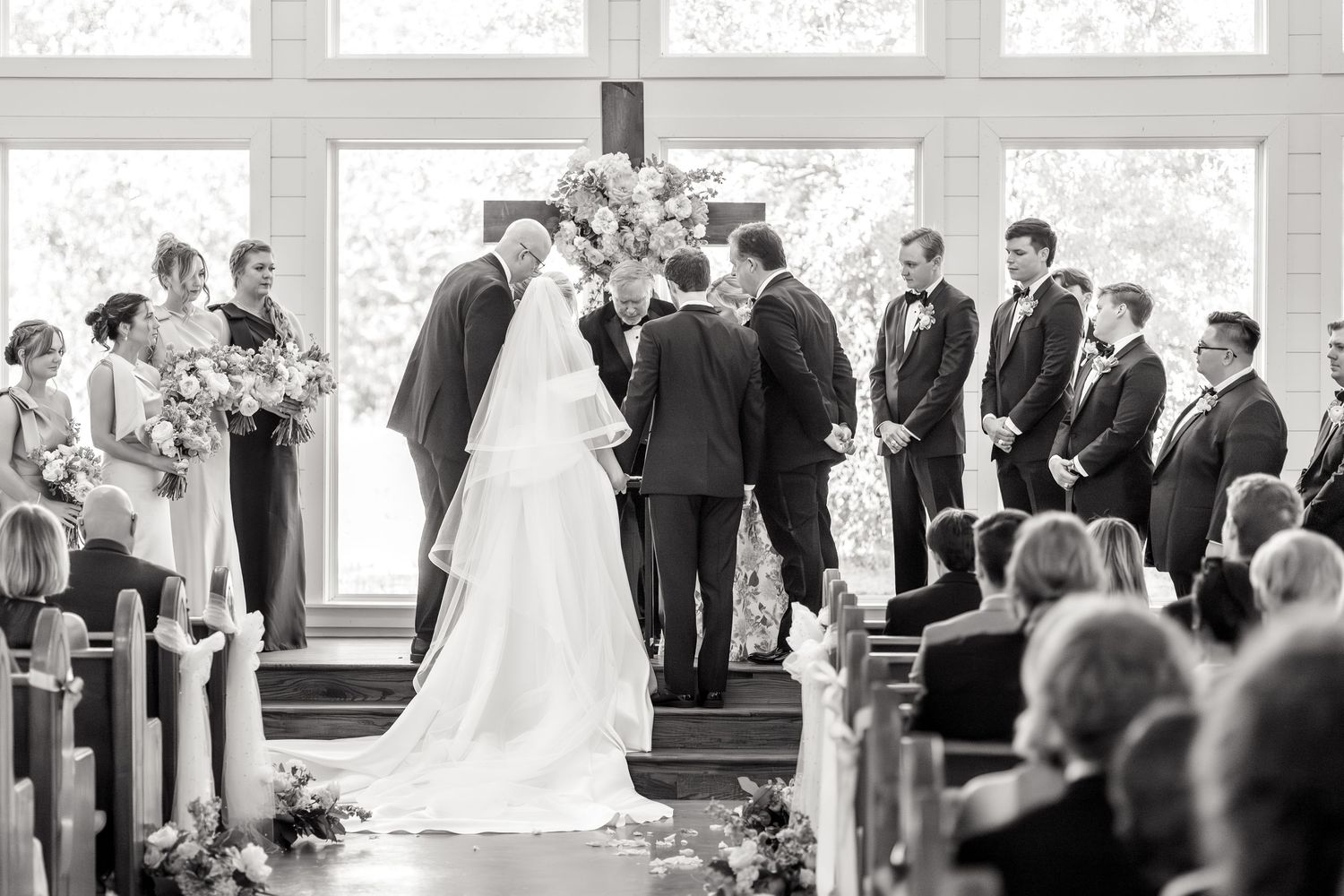 Black and white image of a bride and groom and their parents praying together during Dallas wedding ceremony