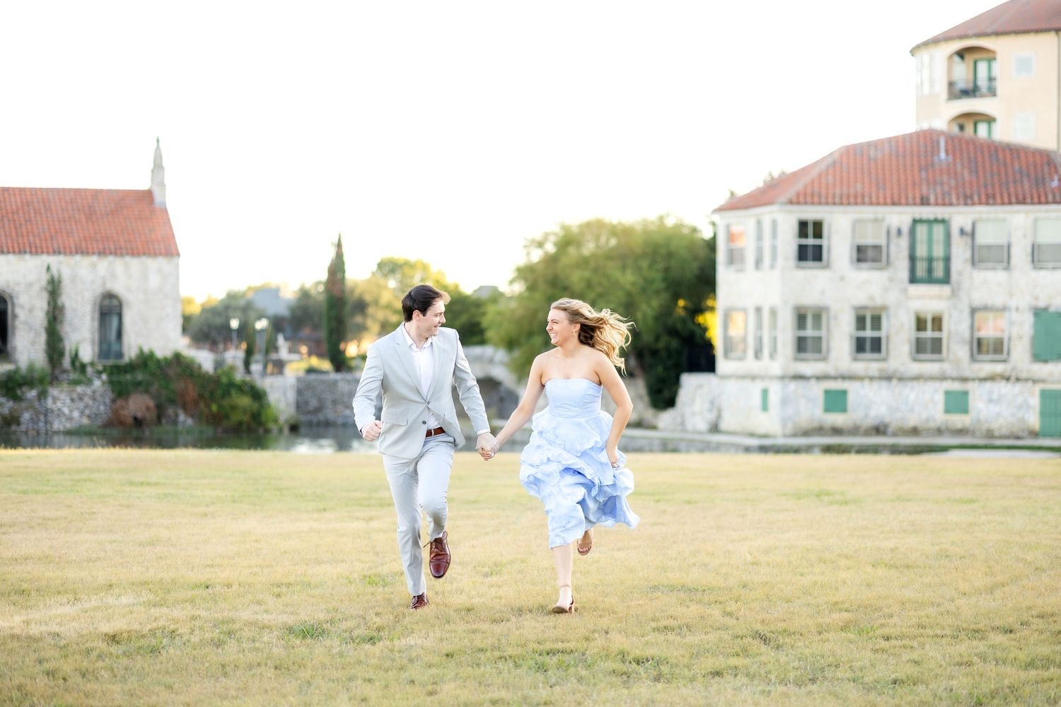 Engaged couple running holding hands in a large field at Adriatica Village in front of a beautiful stone chapel during engagement session