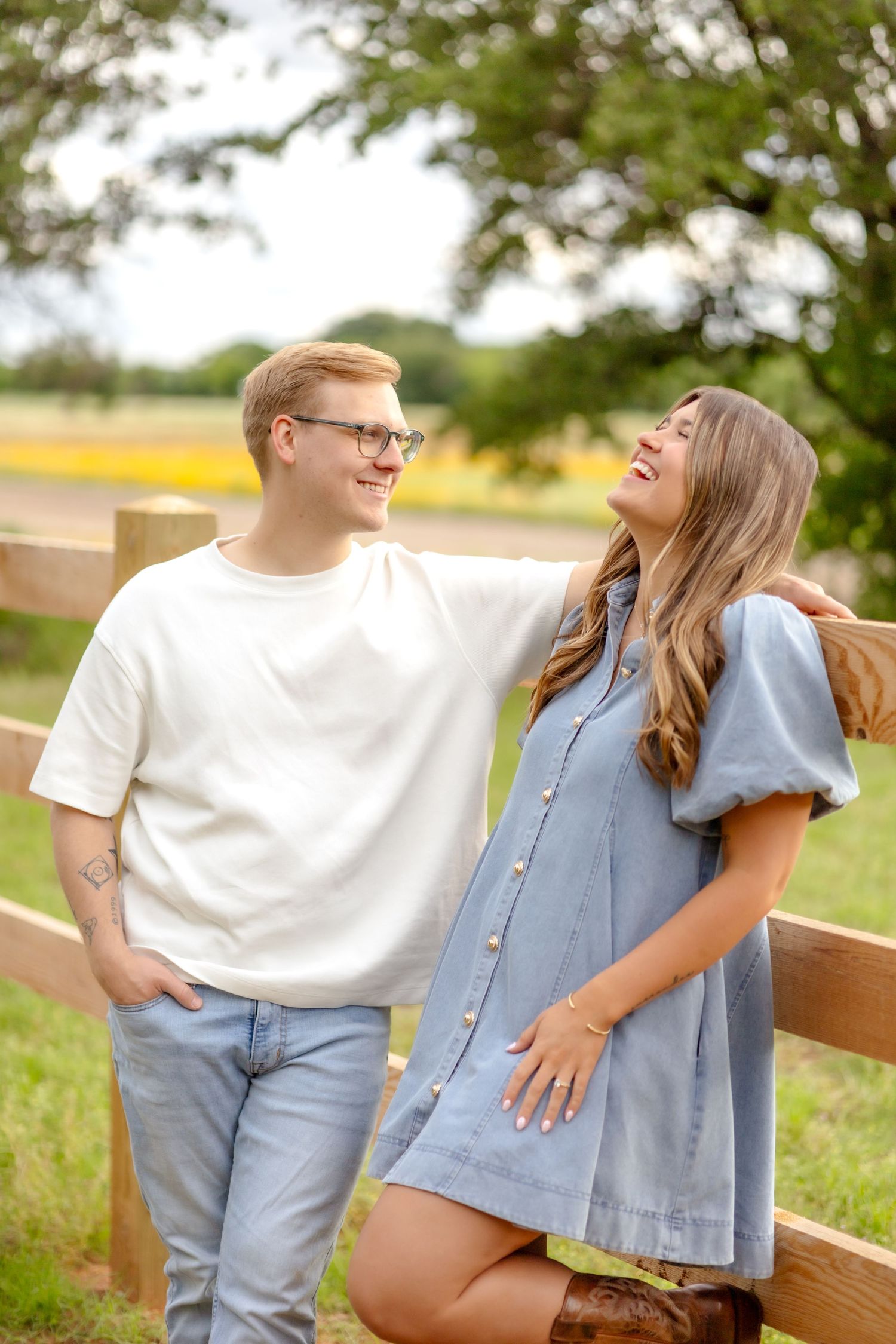 Husband and wife leaning against a wooden fence and him making her laugh during couple's photo session in Abilene, TX