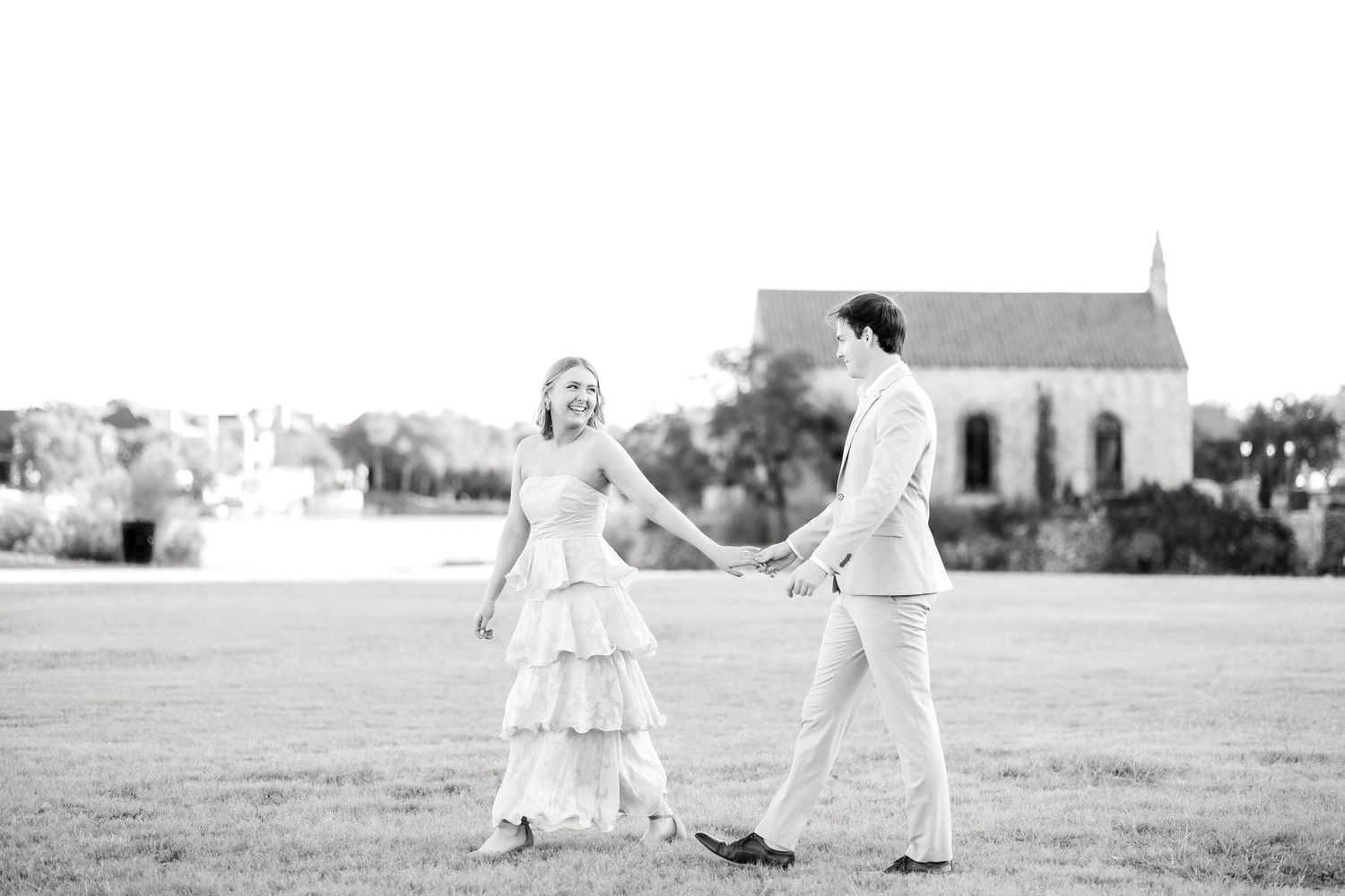 Bride-to-be leading her fiancé walking in a field in front of the chapel at Adriatica Village during Dallas engagement session