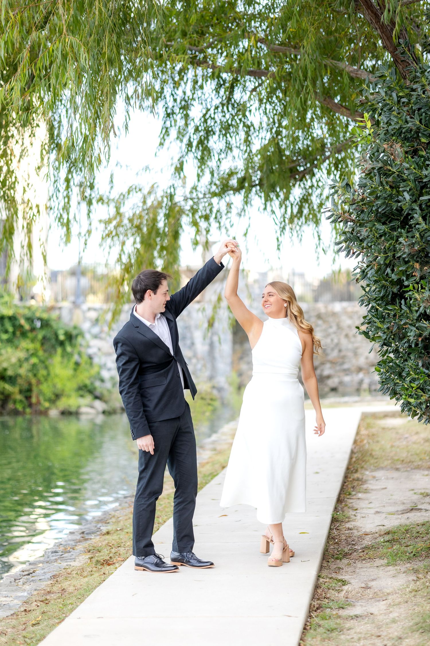 Groom-to-be twirling his fiancé like a princess under a beautiful tree at Dallas engagement session at Adriatica Village