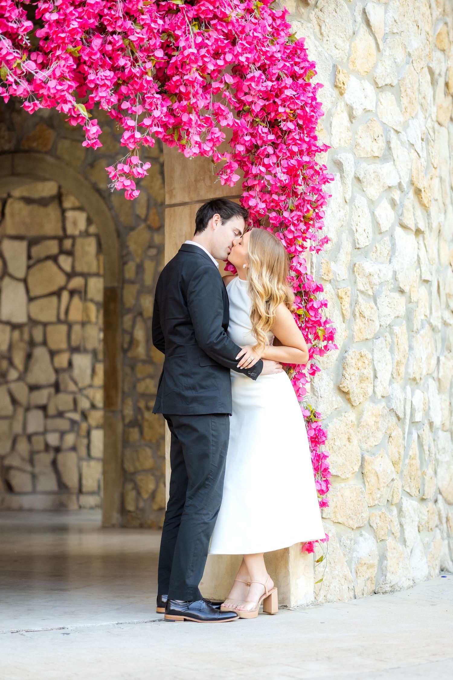 Engaged couple standing against a wall decorated with bright pink florals at Adriatica Village engagement session in Dallas