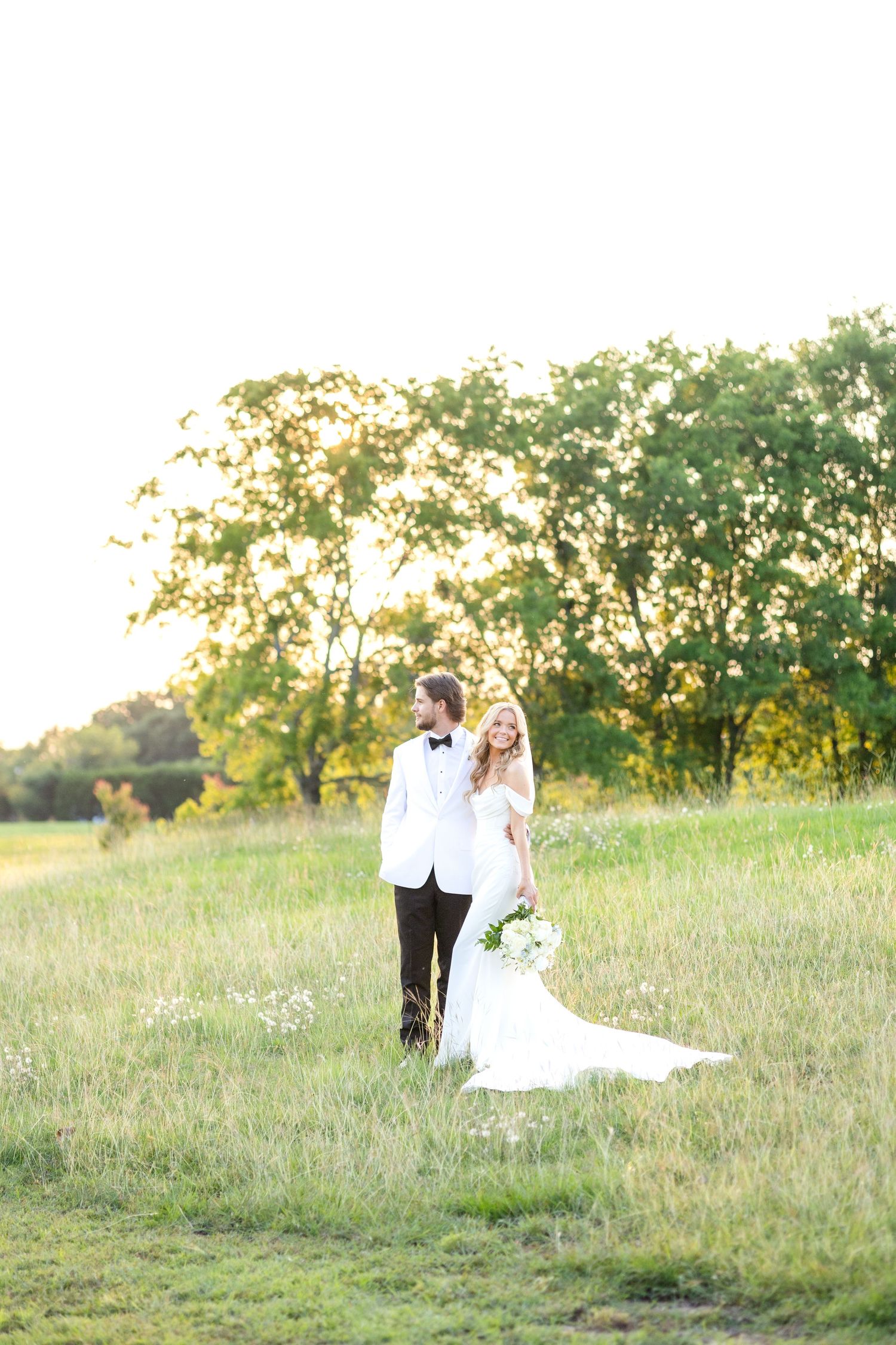 Bride and groom standing together and looking opposite directions in a beautiful field during golden hour at a Dallas wedding venue
