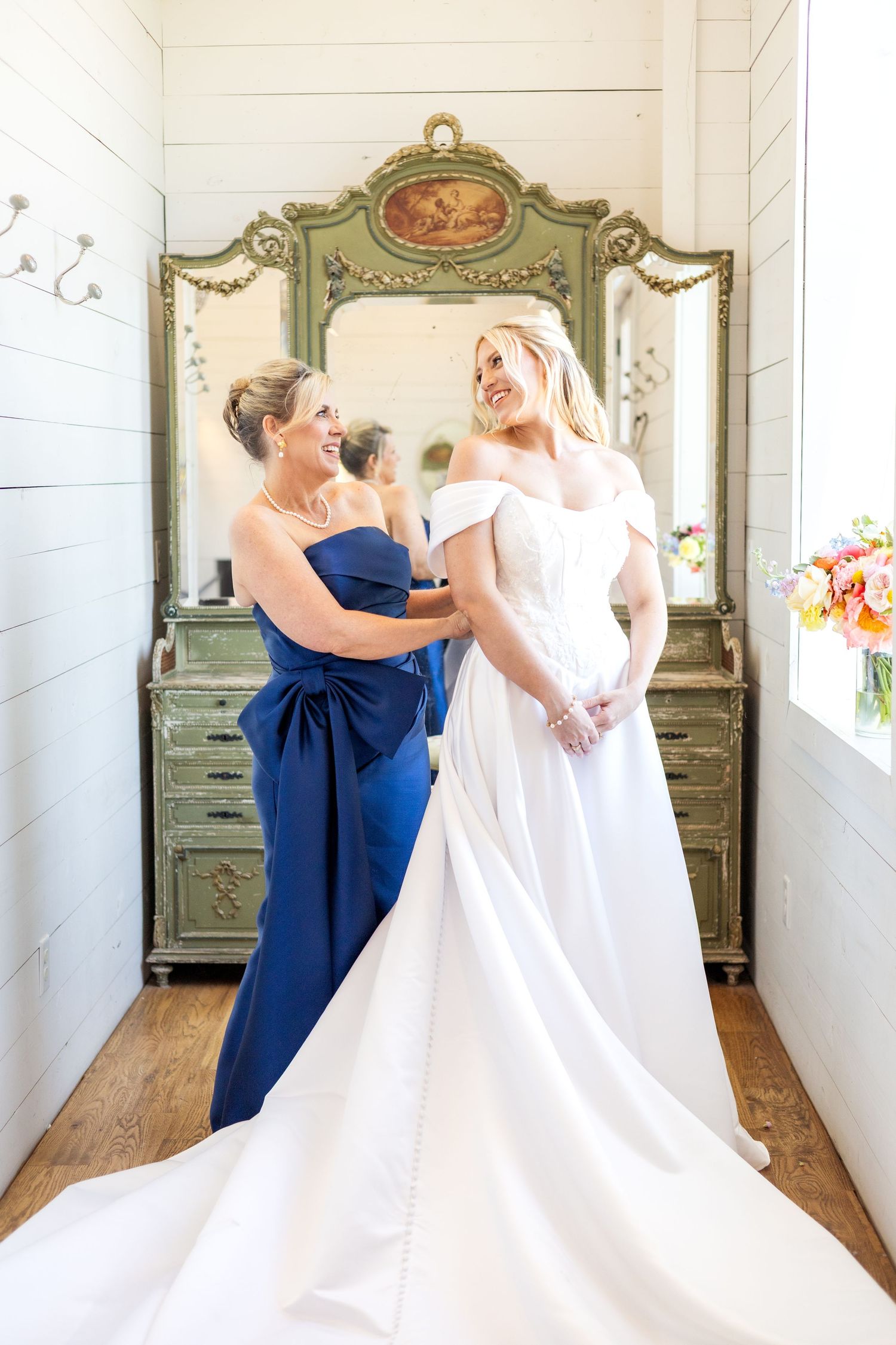 Dallas bride smiling back at her mom as she helps her button up her wedding dress on her wedding day at The Barn at Paradise Place