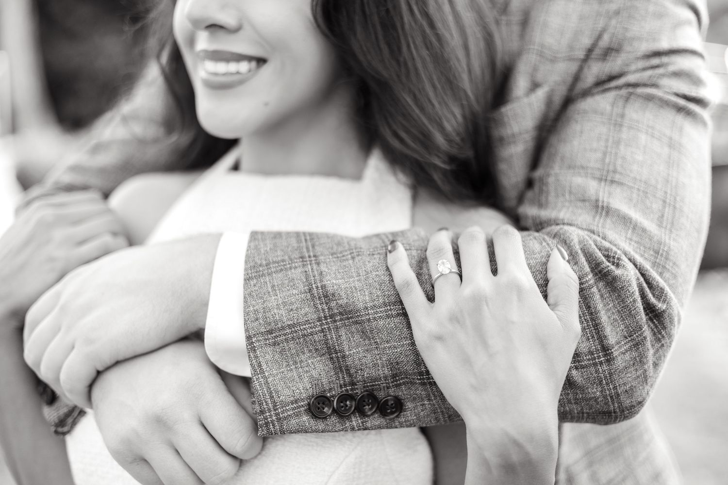 Groom-to-be hugging his fiancé as she rests her hands on his arms and showing her engagement ring during engagement photo session