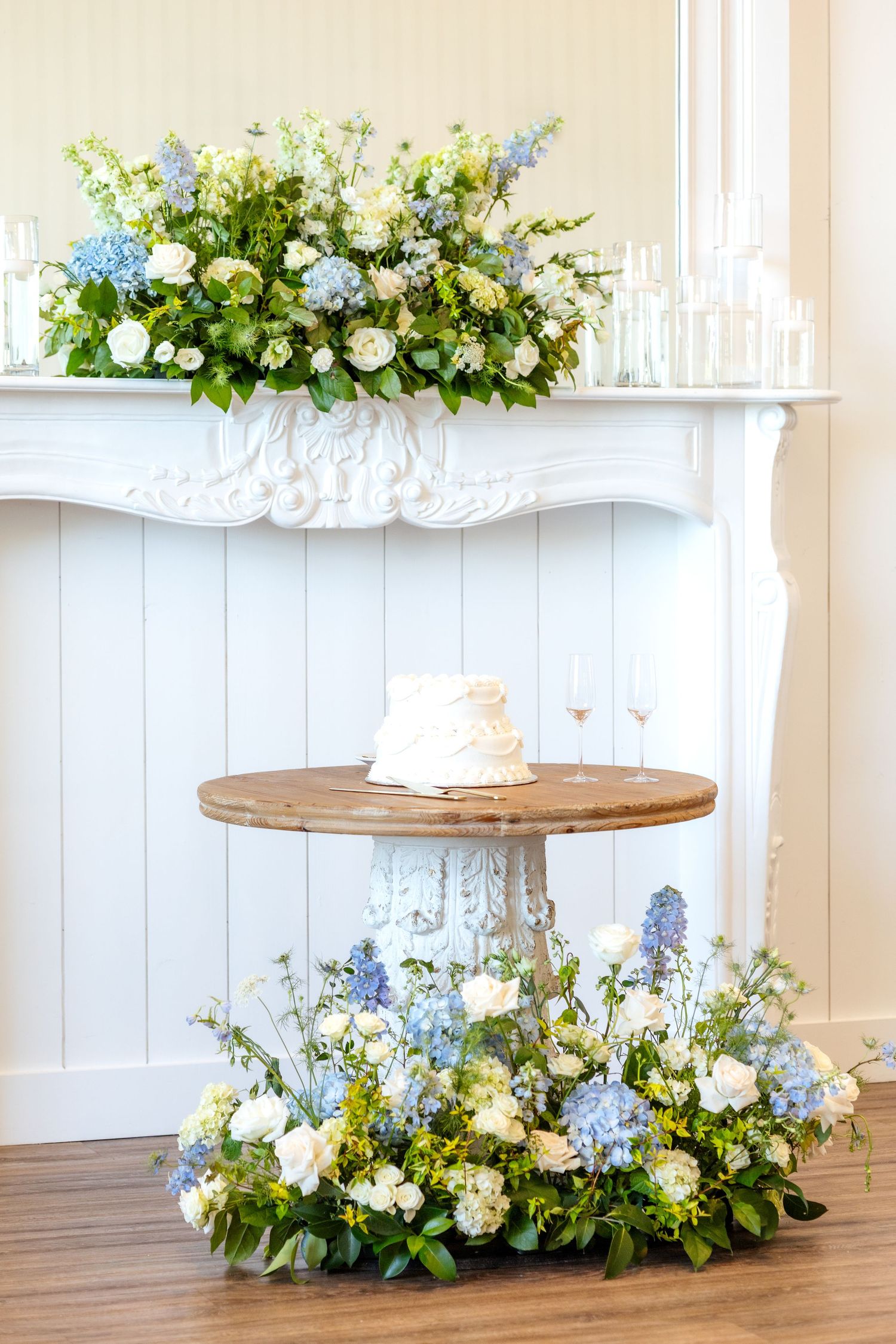 White wedding cake on a table in wedding reception hall at Chandelier Farms surrounded by beautiful Spring florals that are blue and white