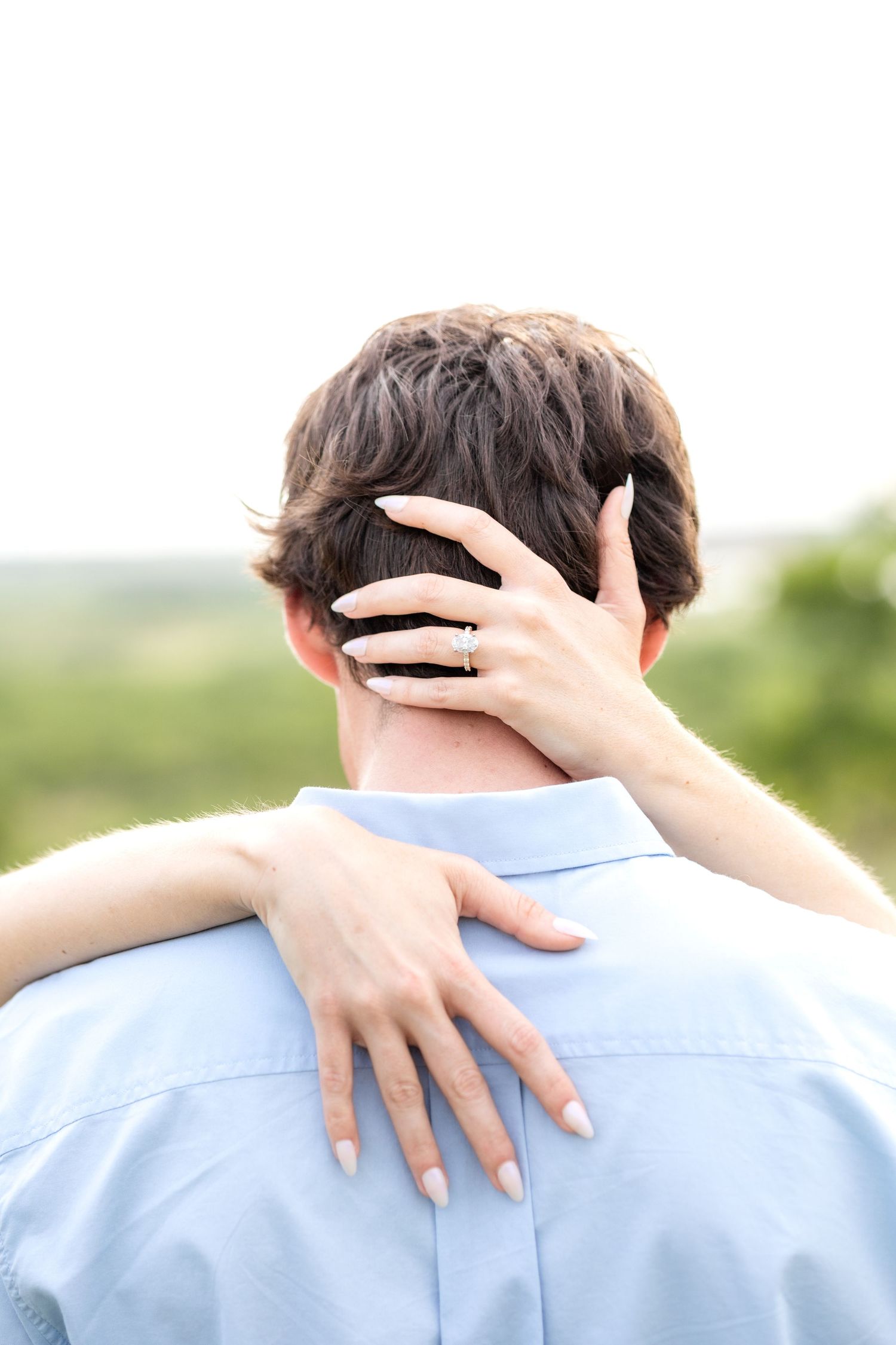 Engaged woman wrapping her arms around her fiancé's back and the back of his head and showing off her engagement ring