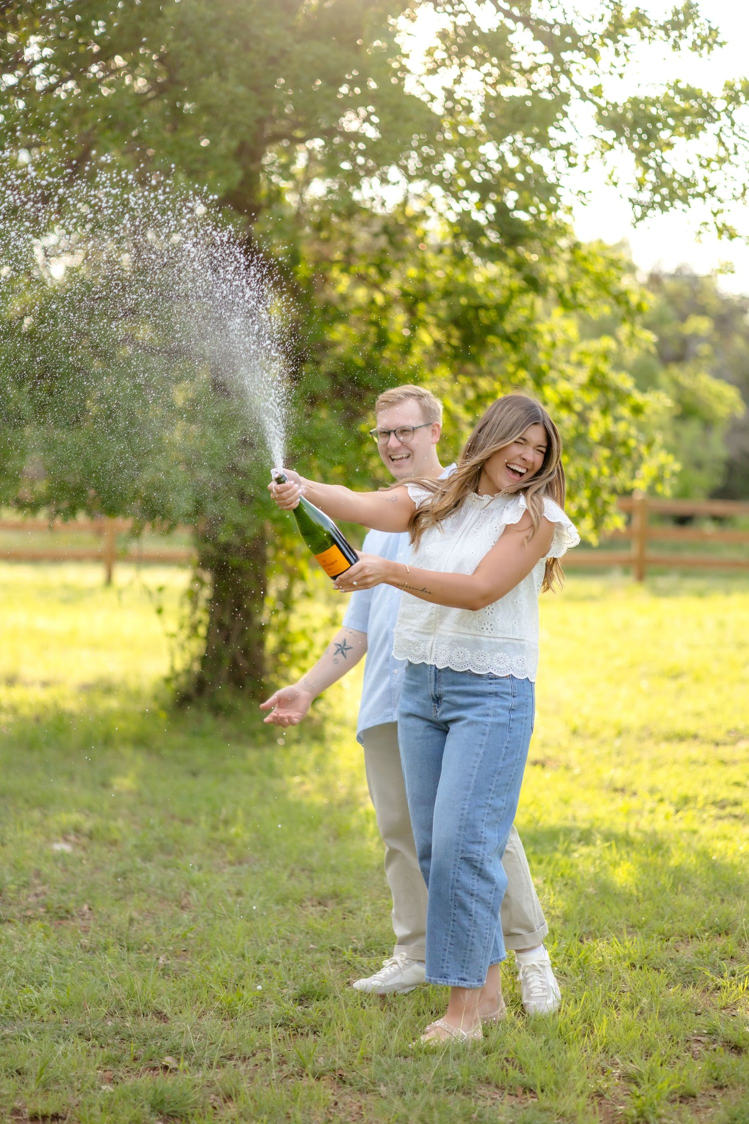 Wife popping a bottle of champagne and the champagne sprays everywhere with her husband standing next to her smiling and laughing