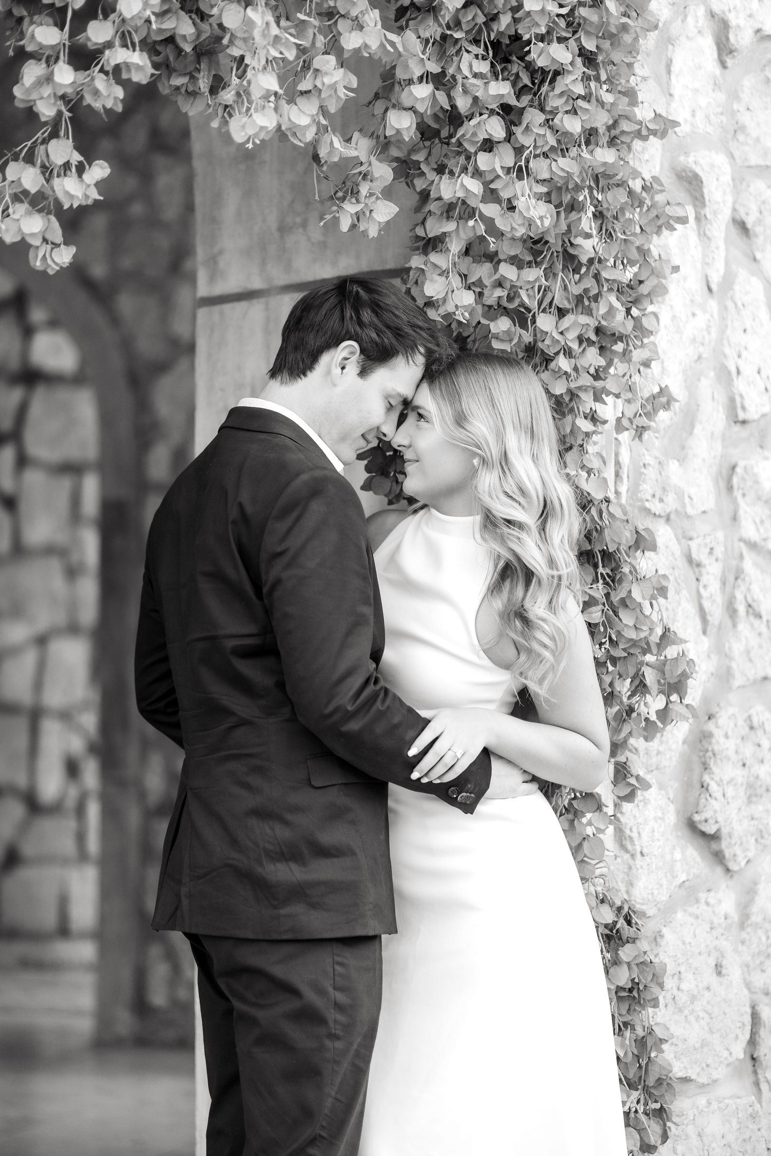 Engaged couple resting foreheads together beneath beautiful flowers hanging above their heads at engagement session at Adriatica Village