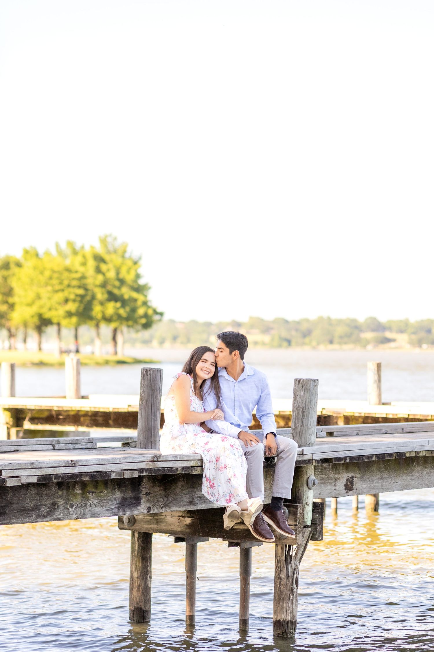 Engaged couple sitting on the side of the dock at White Rock Lake as the groom-to-be kisses his fiancé on the forehead