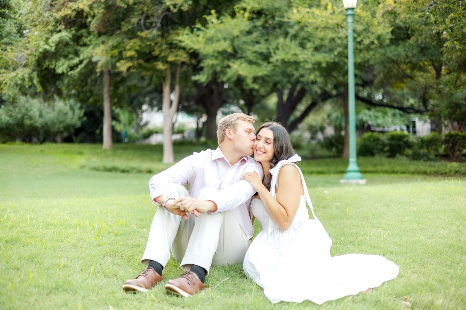 Engaged couple sitting side by side in a field and kissing on the cheek