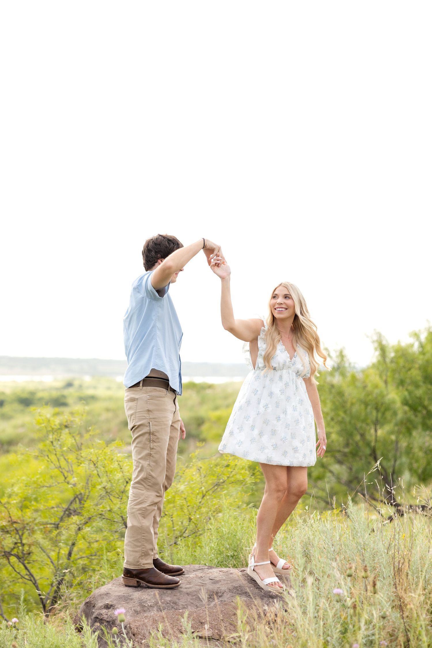 Engaged couple standing on a rock on a Texas ranch and the man spinning his fiancé like a princess at golden hour engagement session