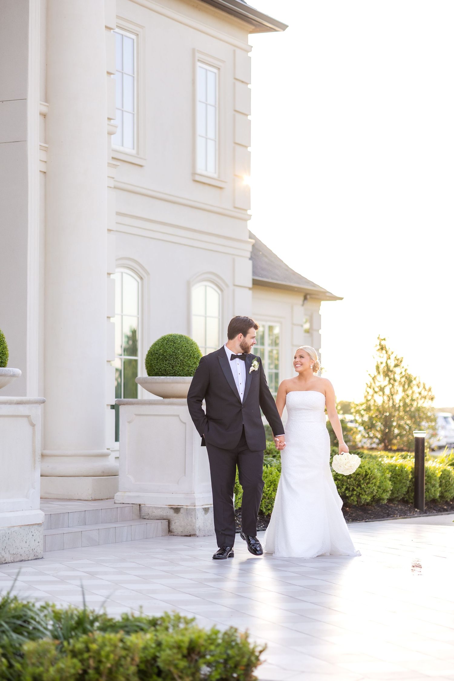 Bride and groom walking and smiling at one another during sunset in front of luxury wedding venue at Dallas summer wedding