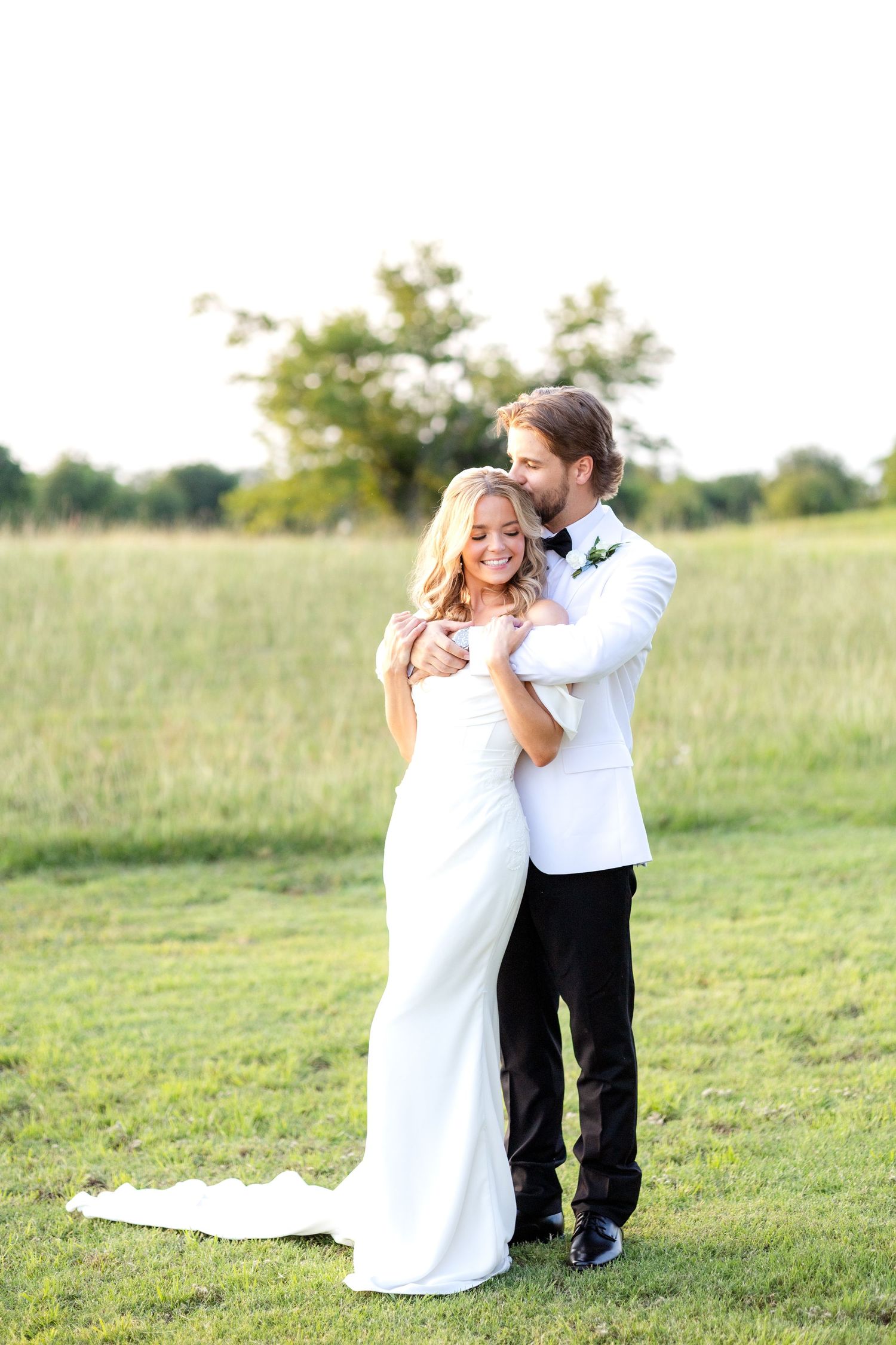 Groom hugging his bride from behind and kissing her on the head in a field at Dallas wedding venue during golden hour