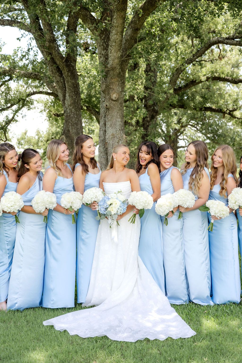 Bride and bridesmaids in light blue dresses holding white bouquets under trees at a Chandelier Farms wedding