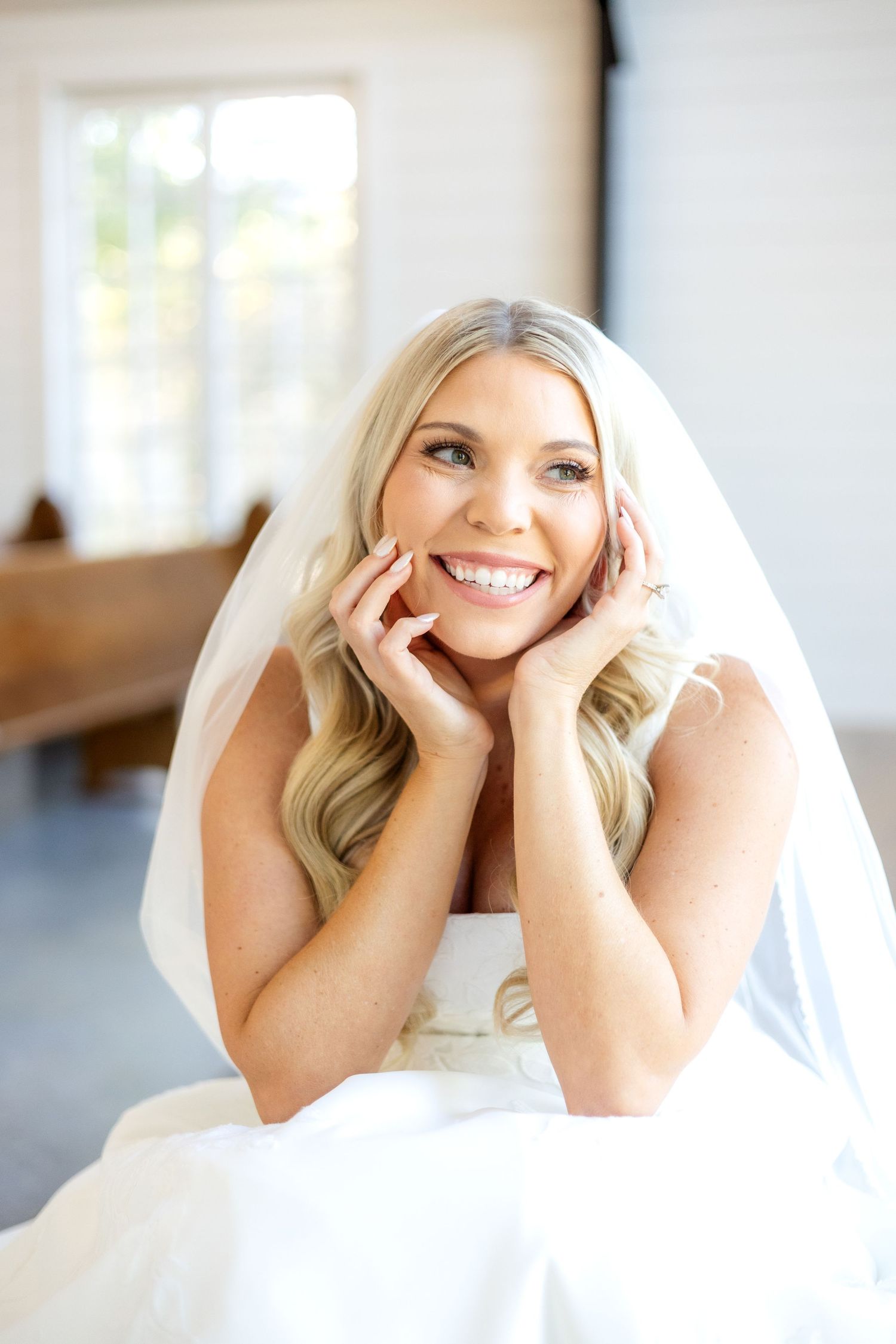Bride resting her head in hands smiling out a window in wedding chapel