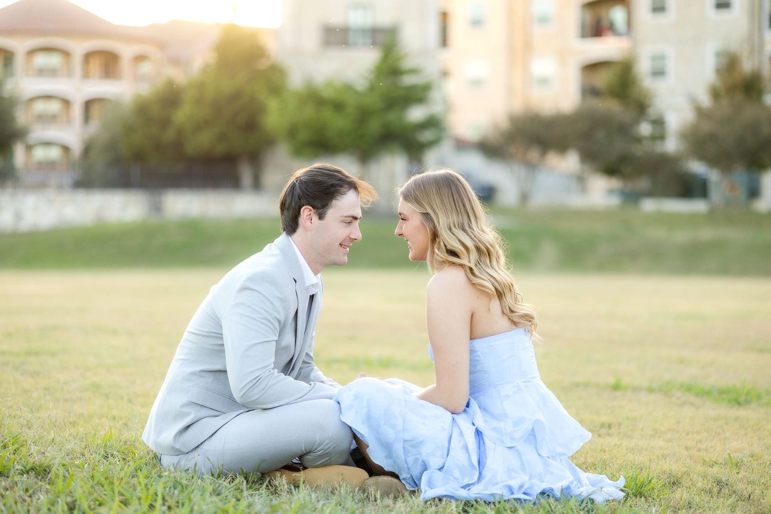 Engaged couple sitting facing one another in a field and holding hands smiling at one another during Dallas engagement session