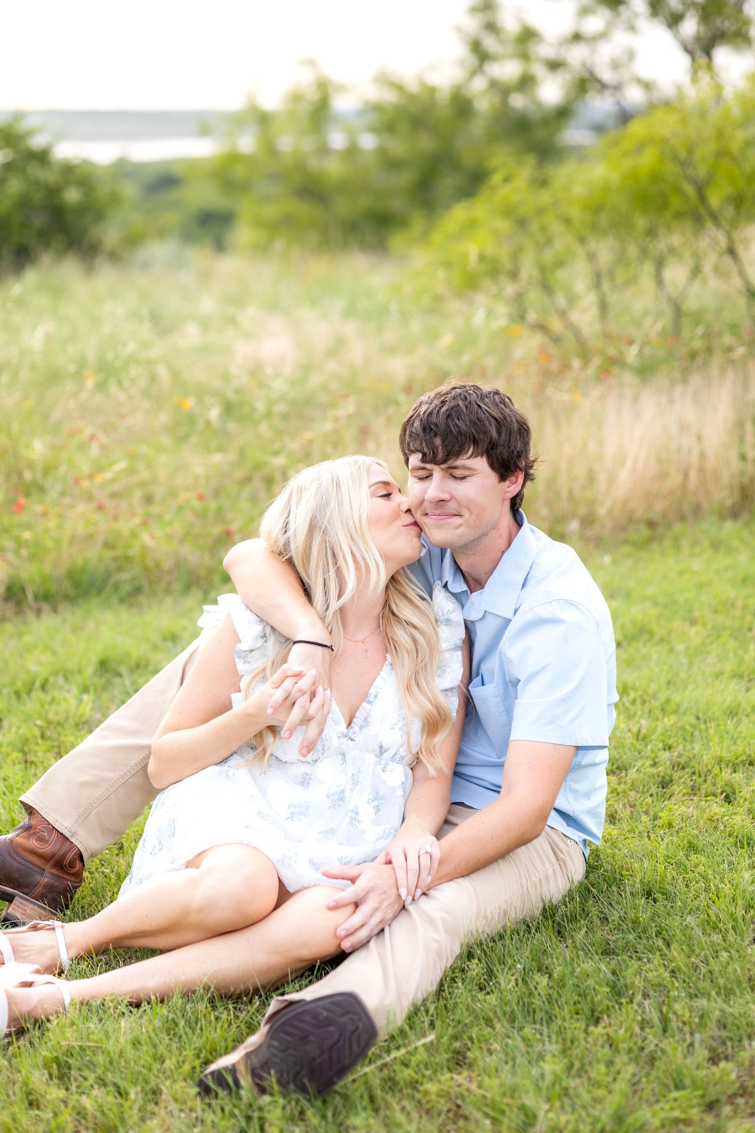 Engaged woman sitting in a field with her fiancé and kissing him on the cheek during dreamy Texas engagement photo session