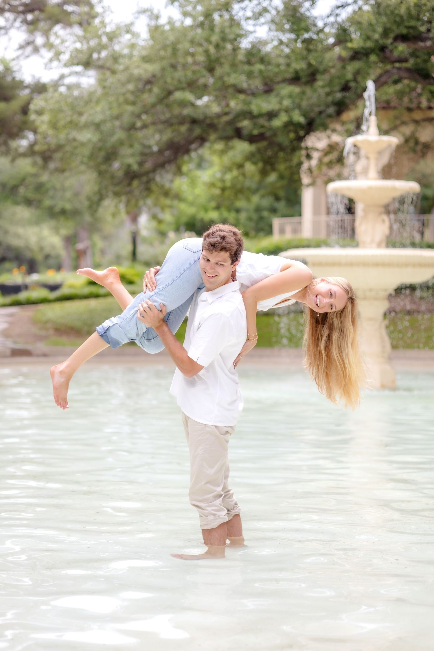 Engaged man lifting his fiancé over his shoulder in a beautiful fountain and spinning around at engagement photo session in Dallas, TX.