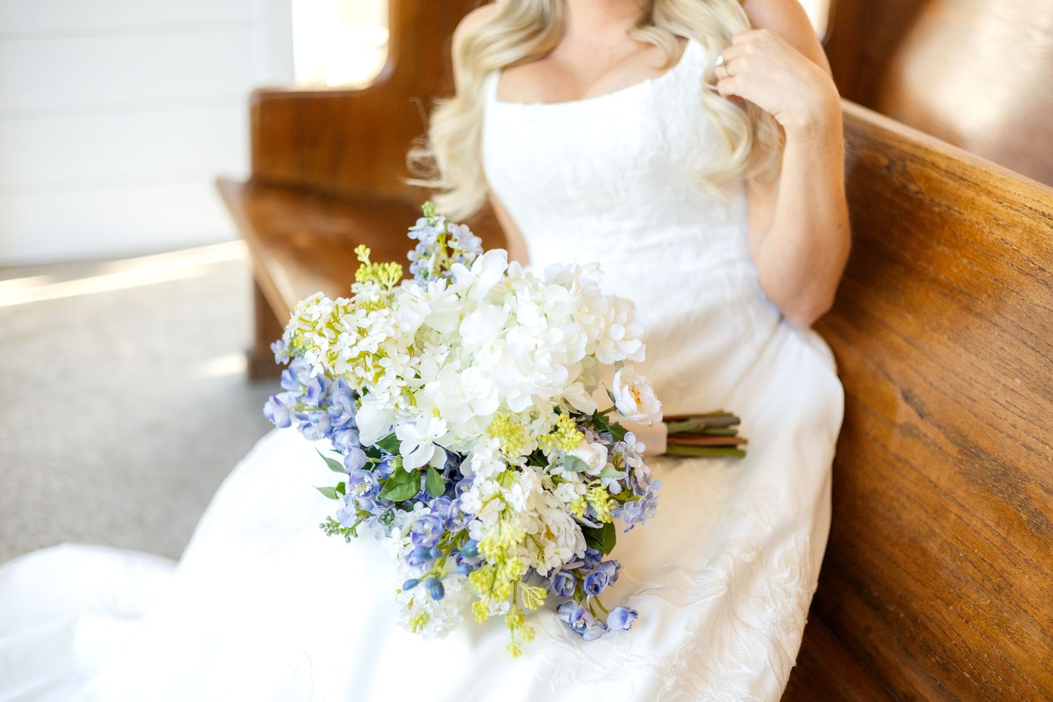 White and blue bridal bouquet resting in Bride's lap while sitting on a wedding chapel pew