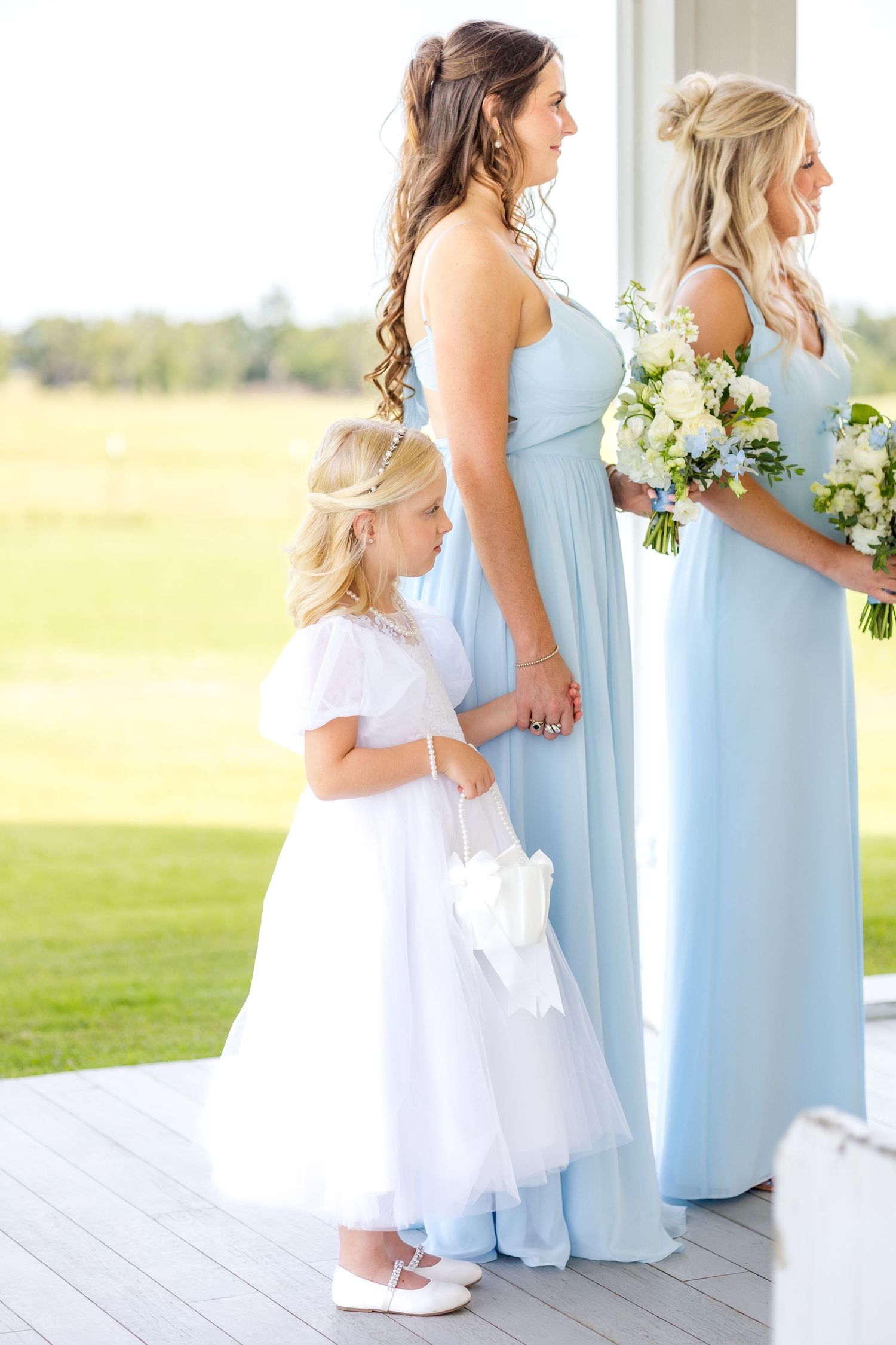 Young flower girl holding a bridesmaid's hand during the wedding ceremony and holding a small white basket with flower petals