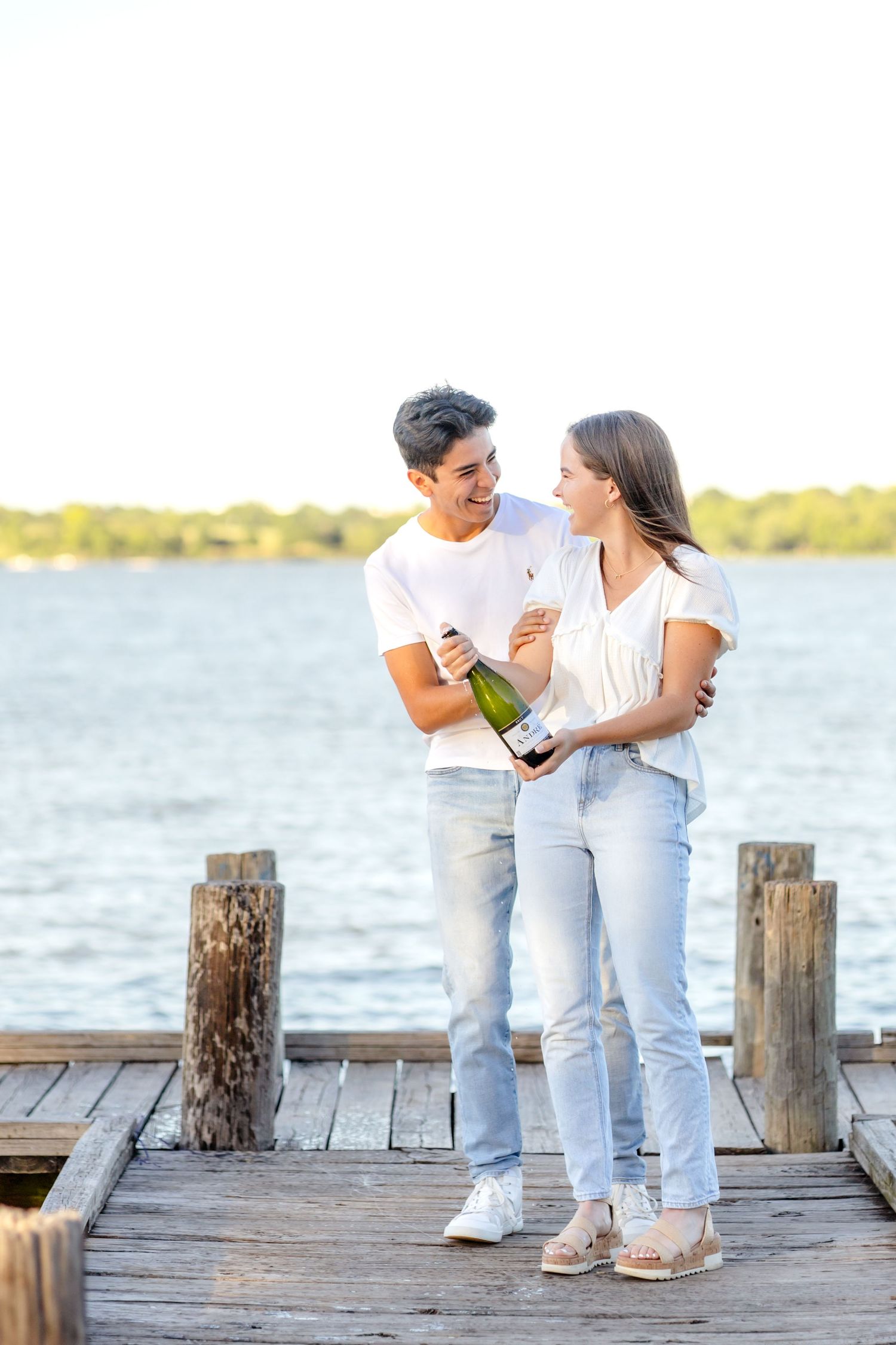 Engaged couple smiling and laughing at one another after popping a bottle of champagne on the dock of White Rock Lake at engagement session