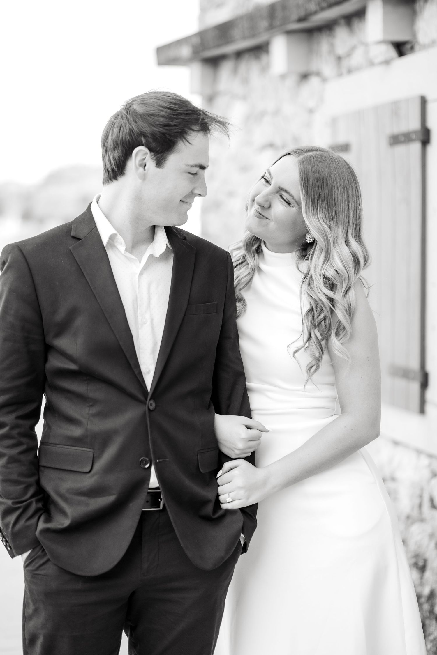 Engaged couple sweetly looking and smiling at one another in front of stone building at Adriatica Village in McKinney, TX