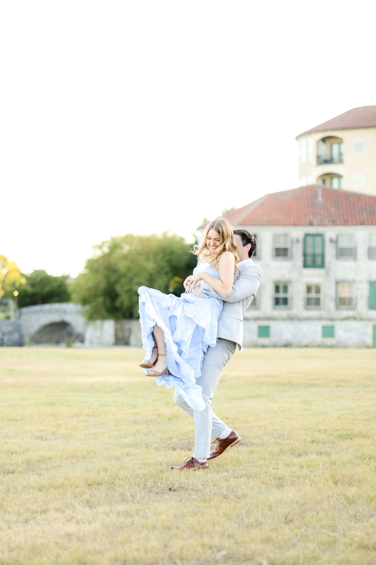 Groom-to-be lifting up his fiancé and spinning her around in a field during engagement session in McKinney, TX at Adriatica Village