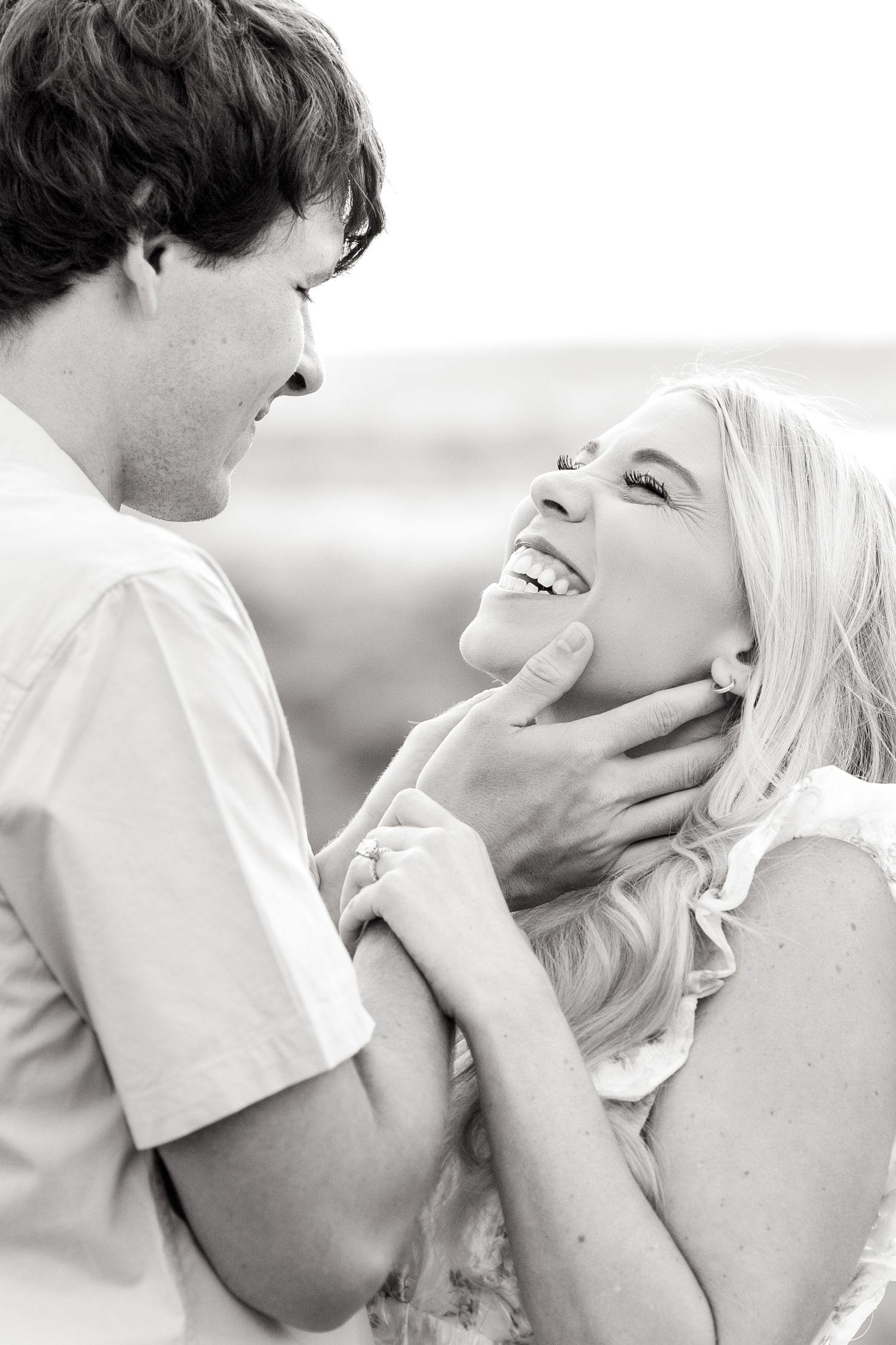Black and white image of engaged man gently holding his fiancé's face as she laughs during Texas engagement photo session