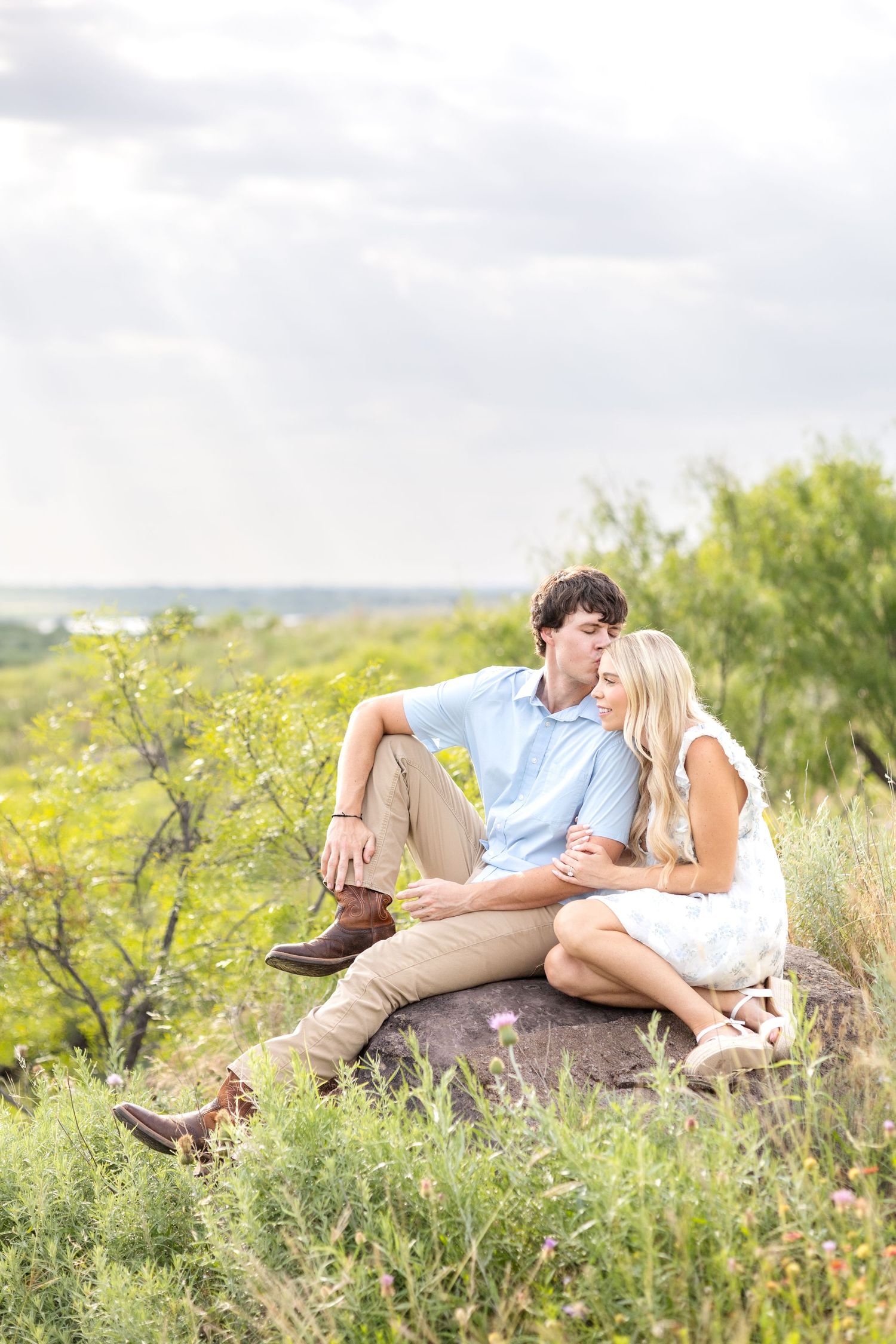 Engaged couple sitting on a rock on a Texas ranch as he kisses his fiancé on her cheek during engagement photo session