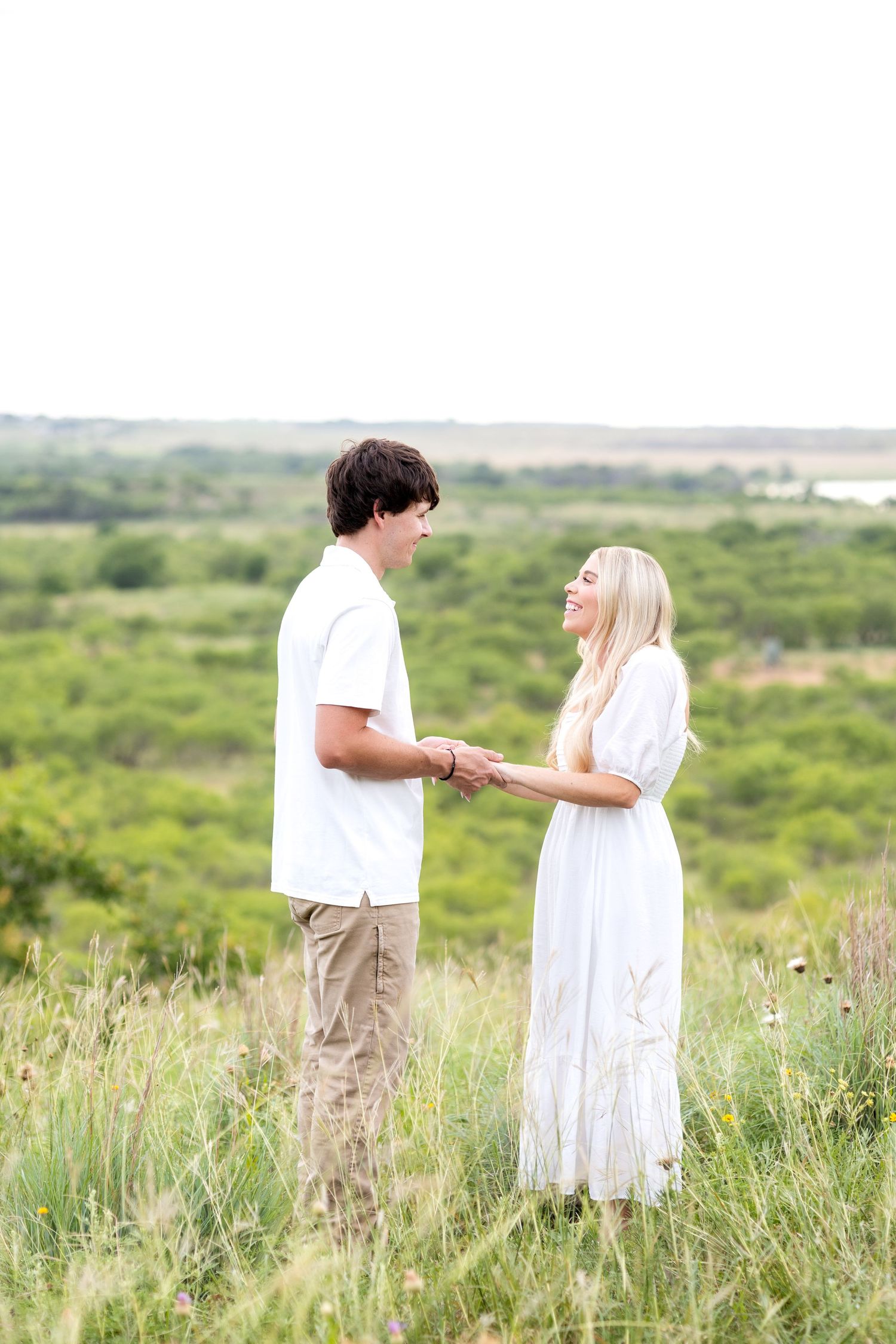 Engaged couple holding hands on a Texas ranch and smiling at one another during sunset engagement photo session