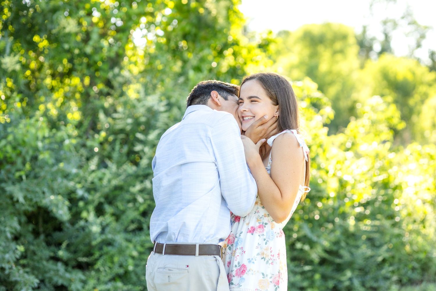 Groom-to-be kissing his fiancé on the cheek as she laughs and smiles in a field at White Rock Lake during engagement photo session