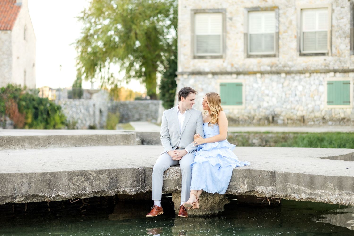 Engaged couple sitting side by side on the dock at Adriatica Village and sweetly smiling at one another during engagement session in Dallas