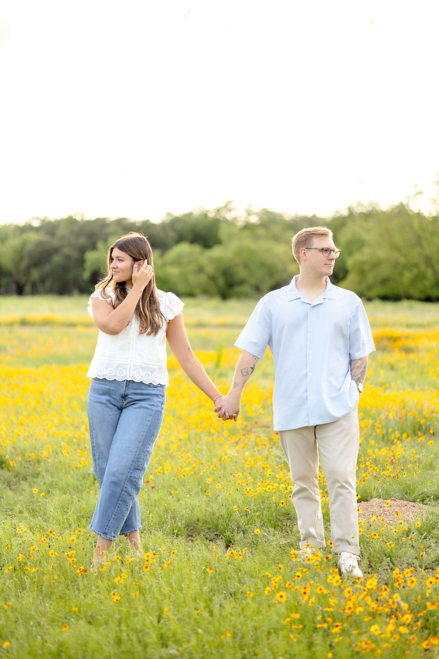 Husband and wife holding hands and looking opposite directions in a wildflower field as she fixes her hair during couple's photo session