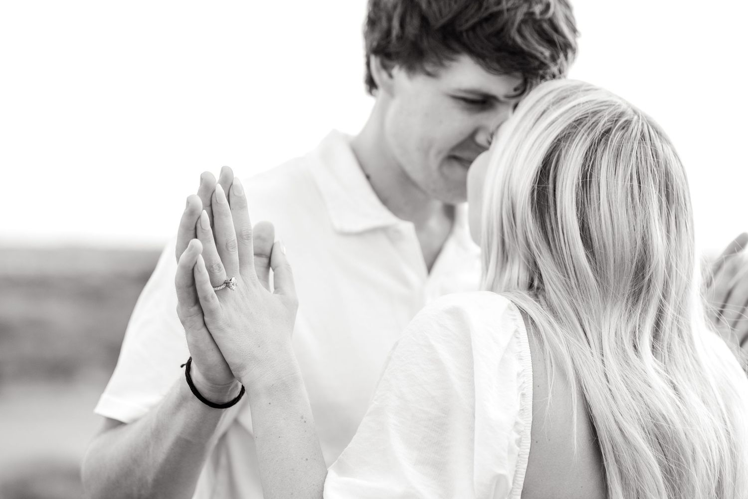 Black and white image of engaged couple going in nose-to-nose and their hands gently touching, showing off the engagement ring
