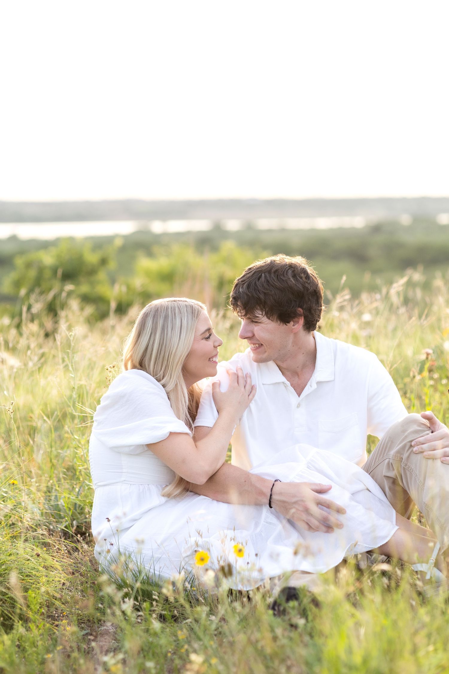 Engaged couple sitting side by side in a golden field in Texas and smiling at one another during engagement photo session