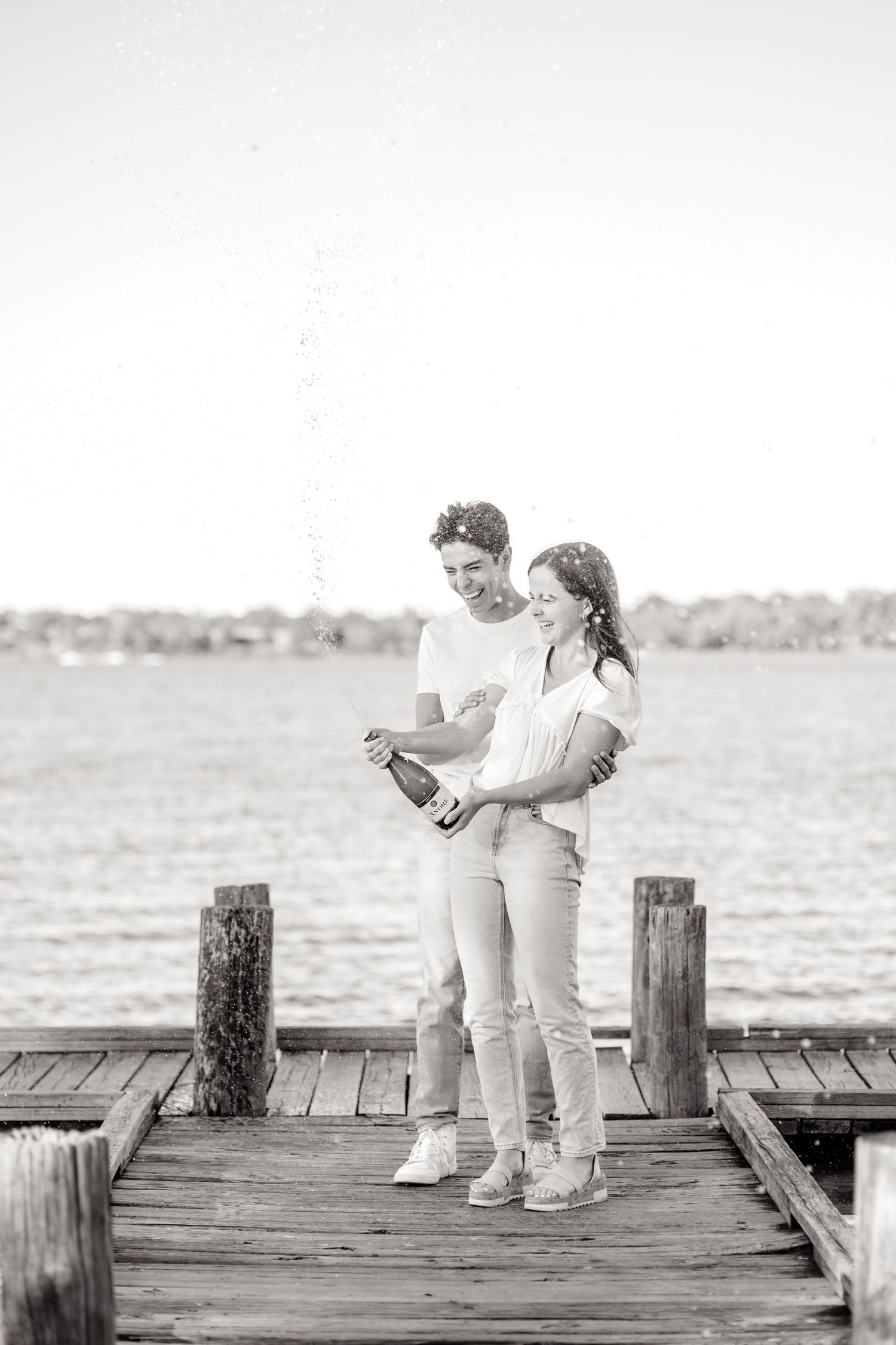 Playful black and white image of an engaged couple popping a bottle of champagne at White Rock Lake during their engagement photo session