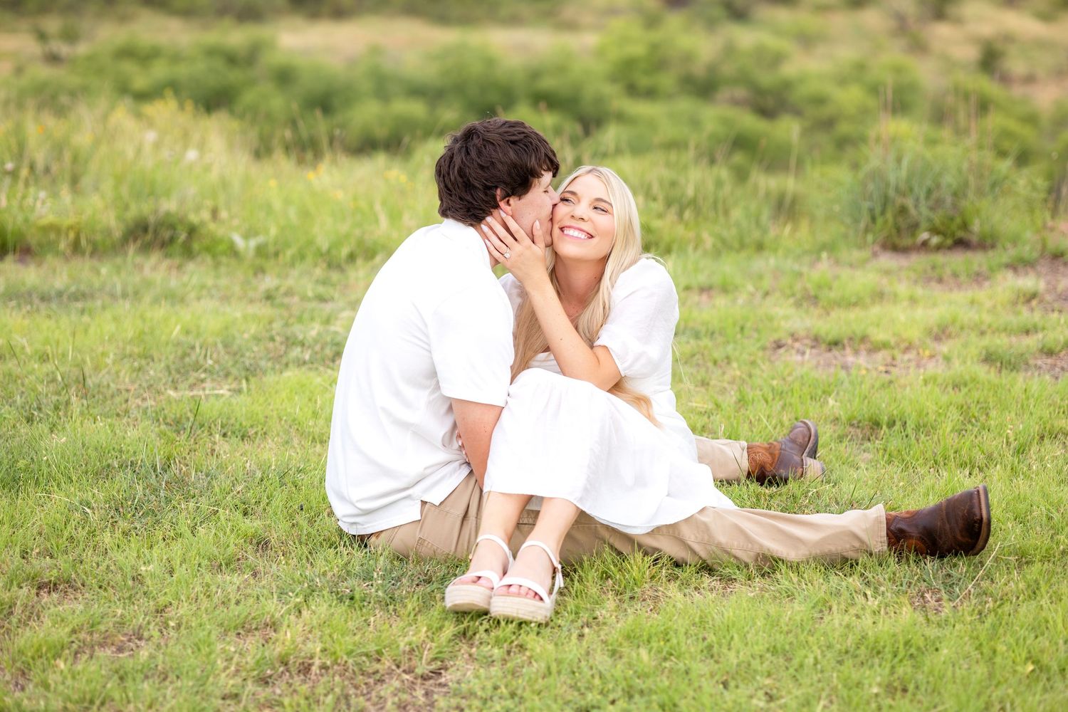 Engaged woman sitting close to her fiance as he gives her a sweet kiss on the cheek at Texas engagement session on a family ranch