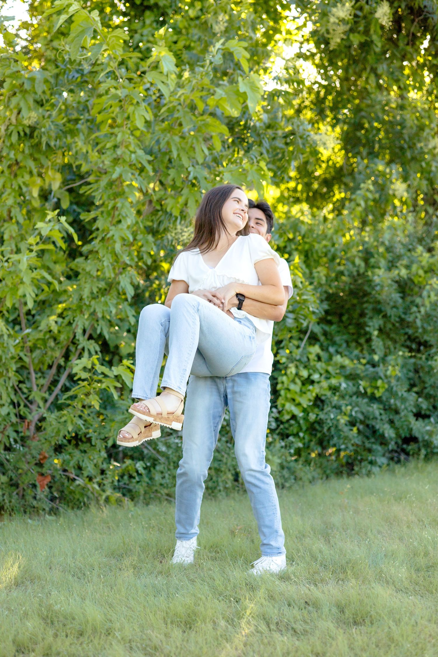 Engaged man picking up his fiancé and spinning around in a field at White Rock Lake during engagement photo session in Dallas
