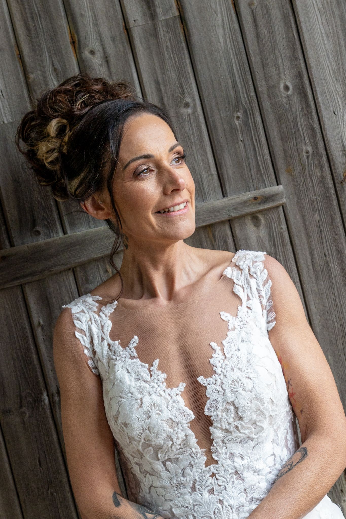 Smiling bride in lace wedding gown with updo hairstyle posing against rustic wooden barn wall.